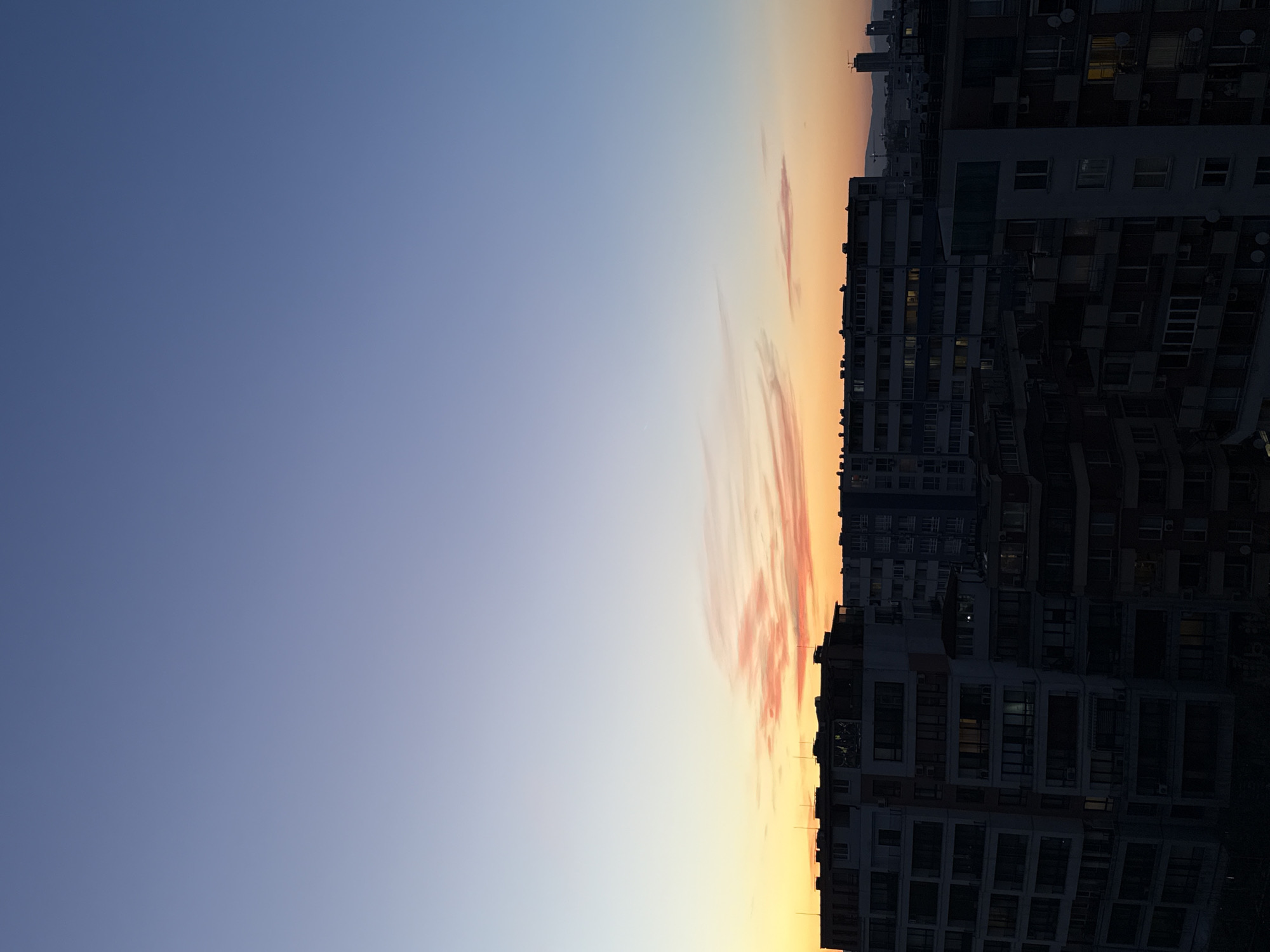 An eye-level, outdoor shot depicts a cityscape at sunset. The sky occupies the left half, transitioning from a deep blue to a soft orange at the horizon. Streaks of pale orange clouds cut through the center. On the right side, multiple apartment buildings are stacked vertically. Many windows are visible, some with lights on inside. The buildings are a dark gray or black. The sky suggests the image was taken in Sofia, Bulgaria at sunset.