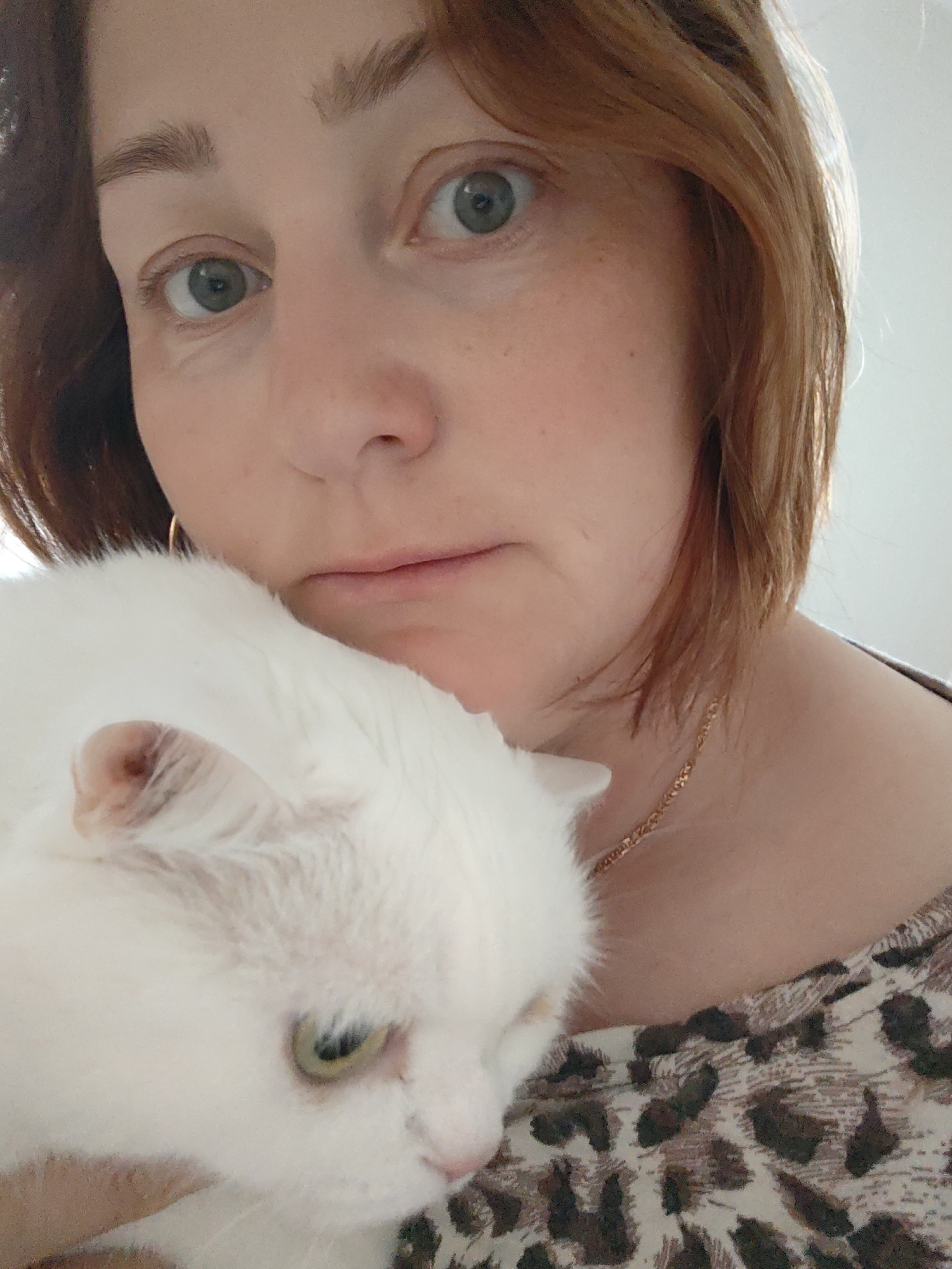 A woman with reddish-brown hair and green eyes is holding a white cat. The woman has fair skin and is wearing gold hoop earrings and a gold necklace. Her shirt has a brown and black animal print. The cat has green eyes and pink ears and nose. The background is blurred and appears to be a light-colored wall. There is no indication in the image that would suggest this was taken in Markovskii, Russia.