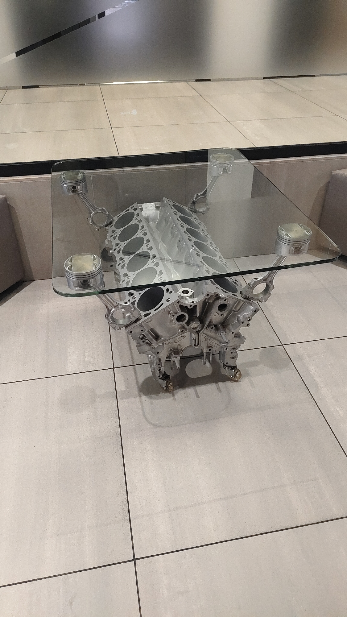 This is a photograph of a unique coffee table made from automotive engine components, set in a modern indoor environment. The main subject is a large, metallic silver engine block, likely a V12 due to the visible cylinder bores, which forms the base of the table. It rests on four small, round feet. A rectangular, clear glass tabletop is supported above the engine block by four polished pistons and their connecting rods, positioned at each corner.
The setting appears to be a clean, contemporary interior space, possibly a lobby, waiting area, or showroom. The floor is covered with large, light-colored square tiles with dark grout lines. In the background, there is a light-toned wall, featuring what looks like a frosted glass panel or a surface with dark horizontal lines, creating a sleek aesthetic. Parts of light-colored, upholstered seating can be seen at the periphery of the image, suggesting a lounge or reception area. No people are visible, and no specific activity is taking place; the image captures a static display. The lighting is consistent with an indoor setting, making the time of day indeterminate. There is some faint, illegible numbering or text embossed on the side of the engine block. The overall impression is one of industrial design repurposed for elegant decor in Krasnoyarsk, Russia.