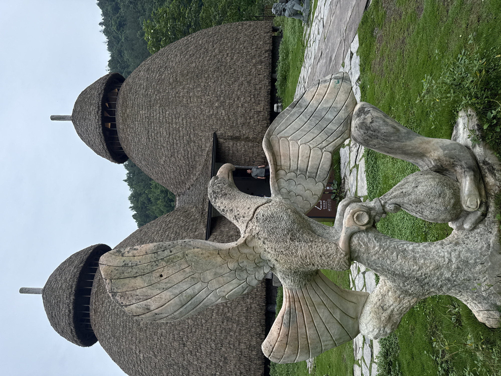 A stone sculpture of a bird with outstretched wings stands in the foreground, its gray stone texture detailed with carved feathers. The sculpture is set against a backdrop of three distinctive thatched-roof structures with rounded tops, likely traditional buildings. A person is partially visible in the doorway of one of the structures. The environment includes stone pathways, green grass, and trees in the background, suggesting a park or cultural site. The location is Jundiping, China.