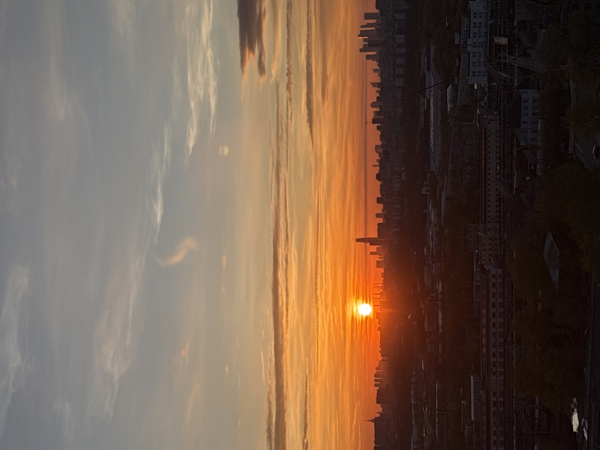 The image shows a cityscape at sunset in Moscow, Russia. A bright orange sun is near the horizon, partially obscured by buildings in the city's skyline. The sky above the sun transitions to lighter hues of orange, yellow, and gray. Thin, streaky clouds are visible in the sky. In the foreground, dark buildings and trees are silhouetted against the bright sky. A bridge and roads can be seen winding through the urban landscape.