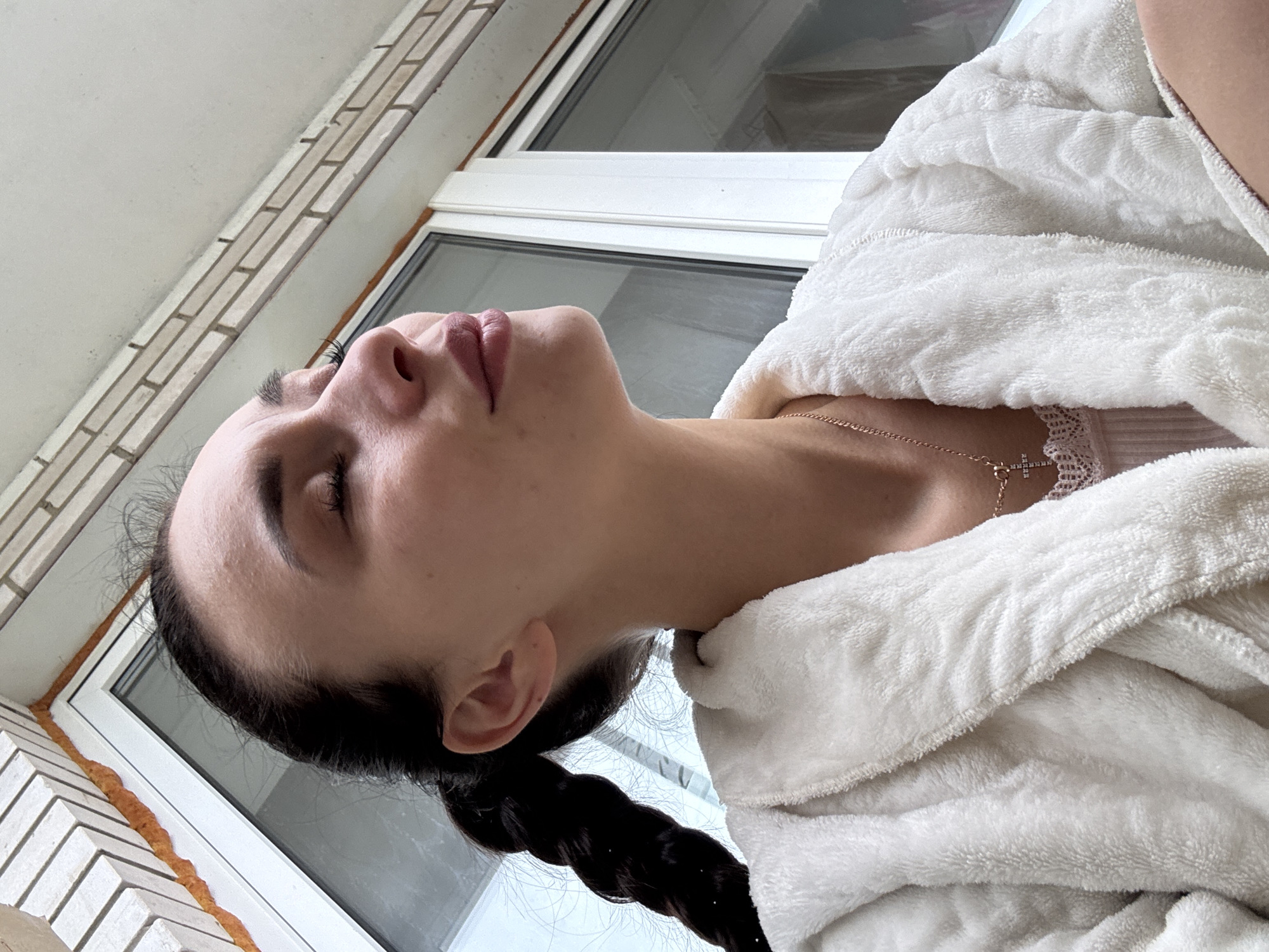 A woman with dark hair pulled back in a braid is wearing a white robe and a delicate necklace with a cross pendant. She is positioned near a window with a white frame. Outside the window is brickwork. The location is Chaikovsky, Russia.