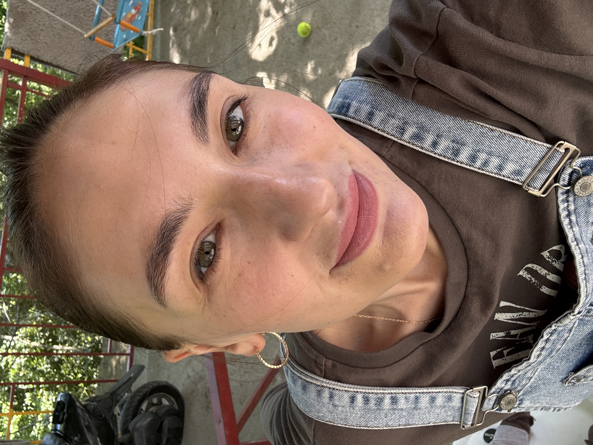 A woman with brown hair pulled back and wearing denim overalls over a brown t-shirt is facing the camera at an upward angle. She has light skin, dark eyebrows, and brown eyes. She wears gold hoop earrings. In the background, there is a tennis ball on concrete, a child's swing set with a blue seat, red and green fencing, and a black moped. The setting appears to be outdoors. It is impossible to confirm if the location is in the city of Sochi, Russia.
