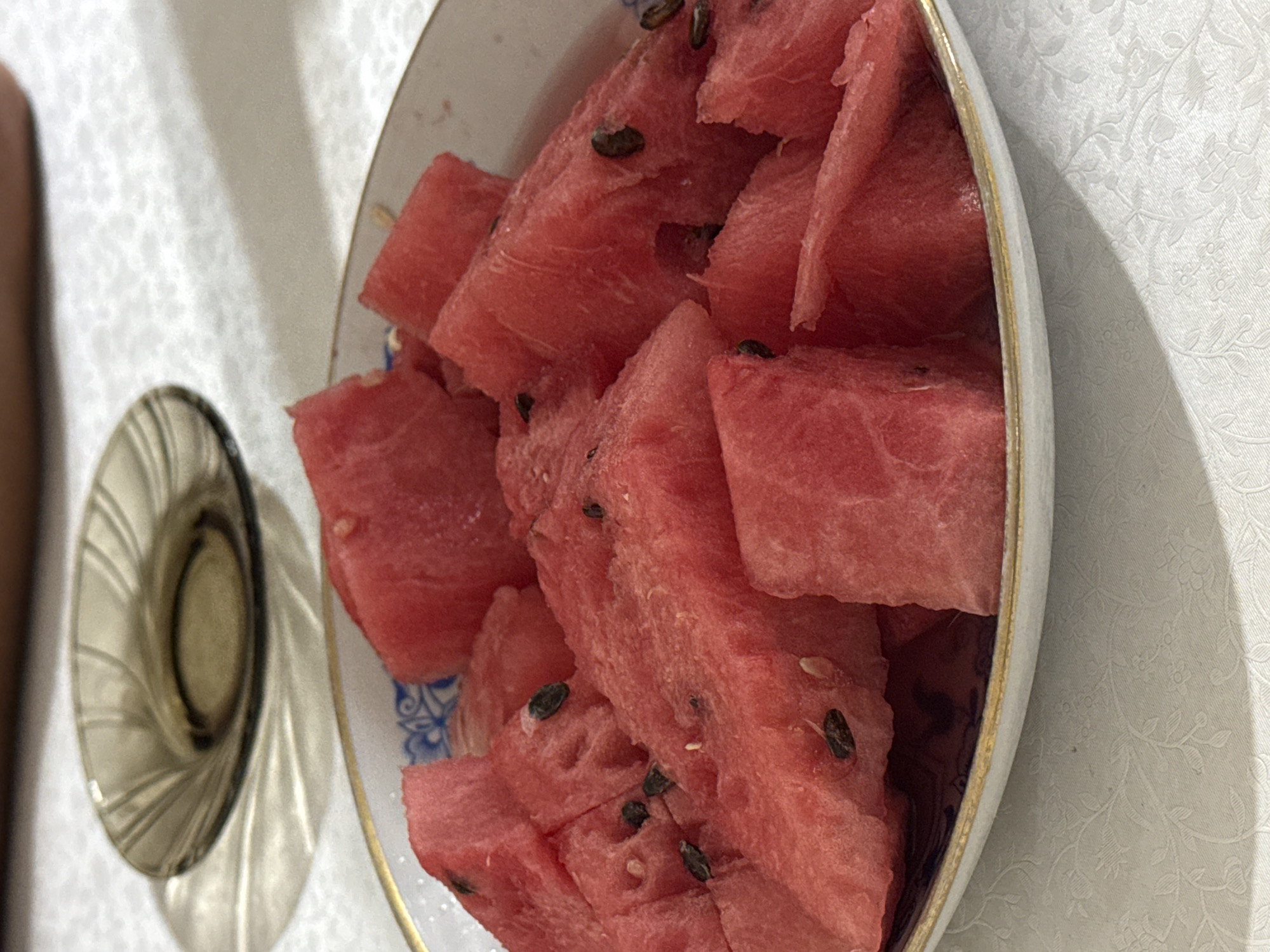 A close-up, overhead shot captures a white ceramic bowl filled with cut pieces of red watermelon. The watermelon slices are irregular in shape, with visible black seeds scattered throughout. The bowl has a decorative blue pattern on its inner rim and a thin gold trim. It rests on a white tablecloth with a subtle floral embossed pattern. To the left of the bowl, a small, dark brown glass dish casts a shadow on the tablecloth. The background shows a blurred human torso and arm. This image was taken indoors, likely in a domestic setting, in the city of Talas, Kyrgyzstan.