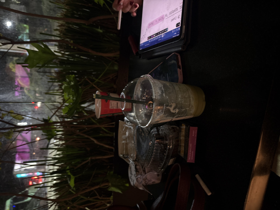 A cluttered dark table hosts a partially consumed green drink in a clear plastic cup with a straw, next to a crumpled paper cup. An iPad displaying a document rests near a phone. Partially visible are purse contents, including a wallet and a small white object. Outside the window, foliage and city lights create a blurred backdrop. A hand holding a cigarette can be seen at the top of the image.