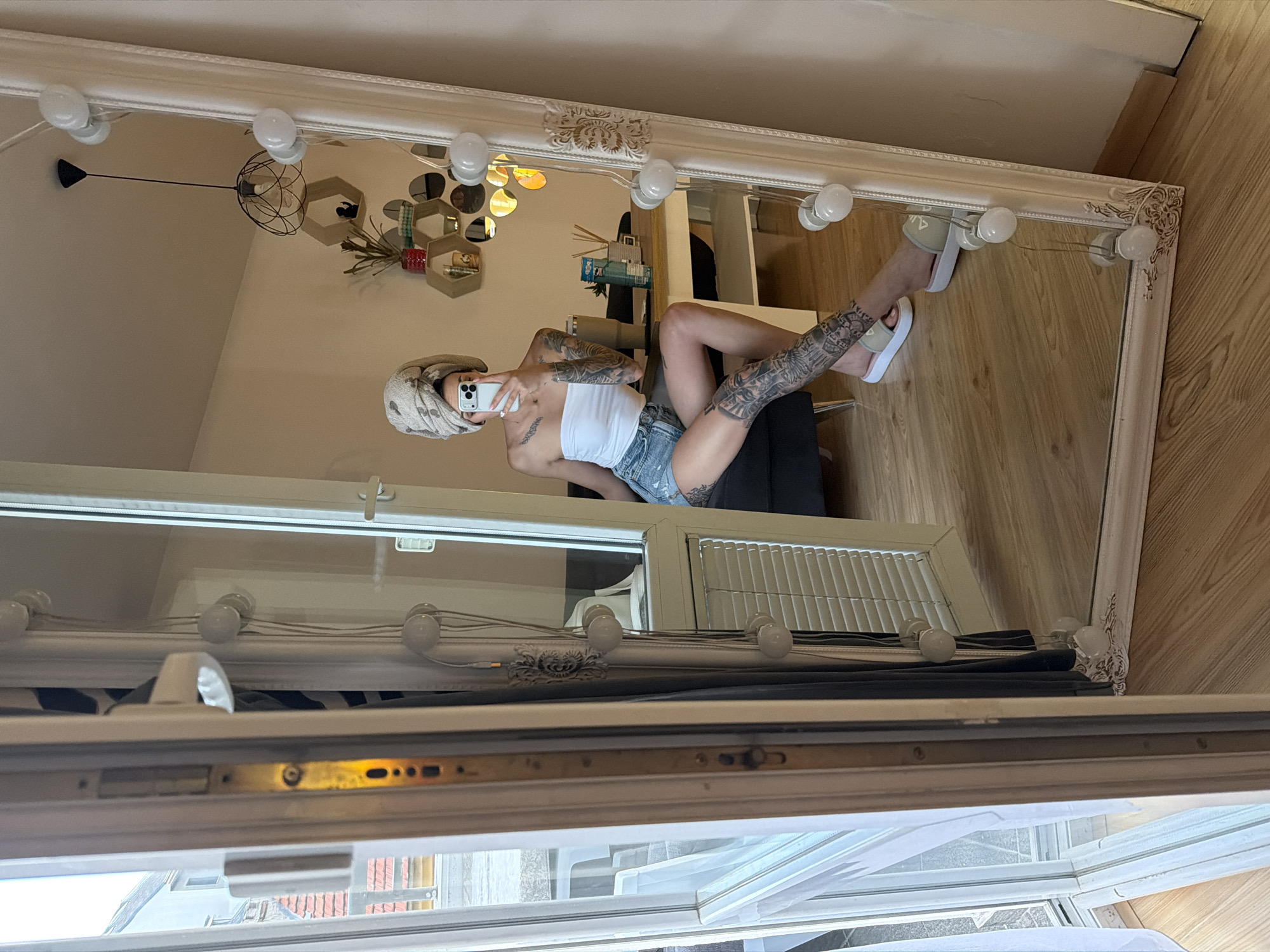 The image captures an indoor scene in Plovdiv, Bulgaria, centered on a full-length mirror reflecting a young adult female taking a selfie. The female is seated on a dark grey sofa, holding a white smartphone (likely an iPhone) at arm's length to photograph her reflection. Her head is wrapped in a light-colored, patterned towel. She wears a white strapless tube top and light blue distressed denim shorts. Prominent dark-ink tattoos cover both her forearms and upper arms, as well as her left leg from the upper thigh to the ankle. She is wearing light-colored slide sandals, one appearing beige and white, the other light green and white.
The large, ornate white-framed mirror, against which a string of approximately ten spherical white vanity lights is draped, leans against a light-colored wall. The reflection in the mirror reveals the room's interior: A light-colored wall adorned with a cluster of hexagonal wooden shelves, some containing items such as a small plant in a red pot, and a collection of circular and irregular-shaped decorative mirrors. A black pendant light fixture with a geometric cage design hangs from the ceiling. A light-toned table with white legs is visible behind the sofa, holding a large grey tumbler, a clear glass bottle with a blue label, and a reed diffuser set. The floor is composed of light wooden floorboards.
To the left of the mirror, a white PVC-framed balcony door or large window is visible. Through this opening, an outdoor scene is discernible, featuring concrete or tiled ground, parts of distant buildings with red-tiled roofs, and other structures under a bright daytime sky. A sheer white curtain hangs to the immediate left of the mirror frame, and a darker curtain along with white horizontal blinds are visible within the reflected window area and to the right of the physical door frame.