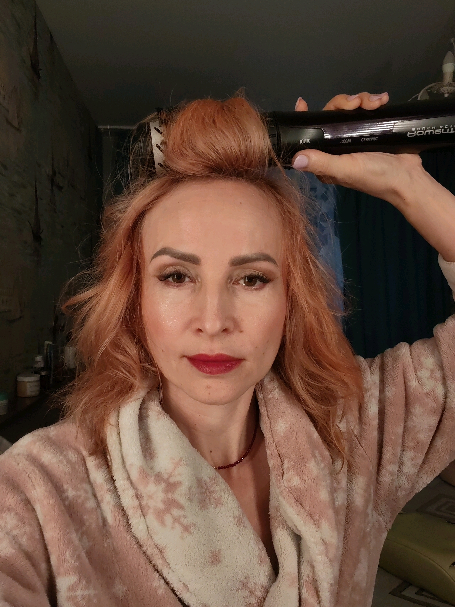 A woman with reddish-blonde hair and makeup, wearing a pink and white patterned robe, holds a black hair dryer with a brush attachment to her hair, which is partially styled into a bun. She is indoors, seemingly in a domestic setting, with a dark wall and a curtain visible in the background. The scene appears to be a selfie taken by the woman. There is no specific information in the image to indicate its location in the city of Chaikovsky, Russia.