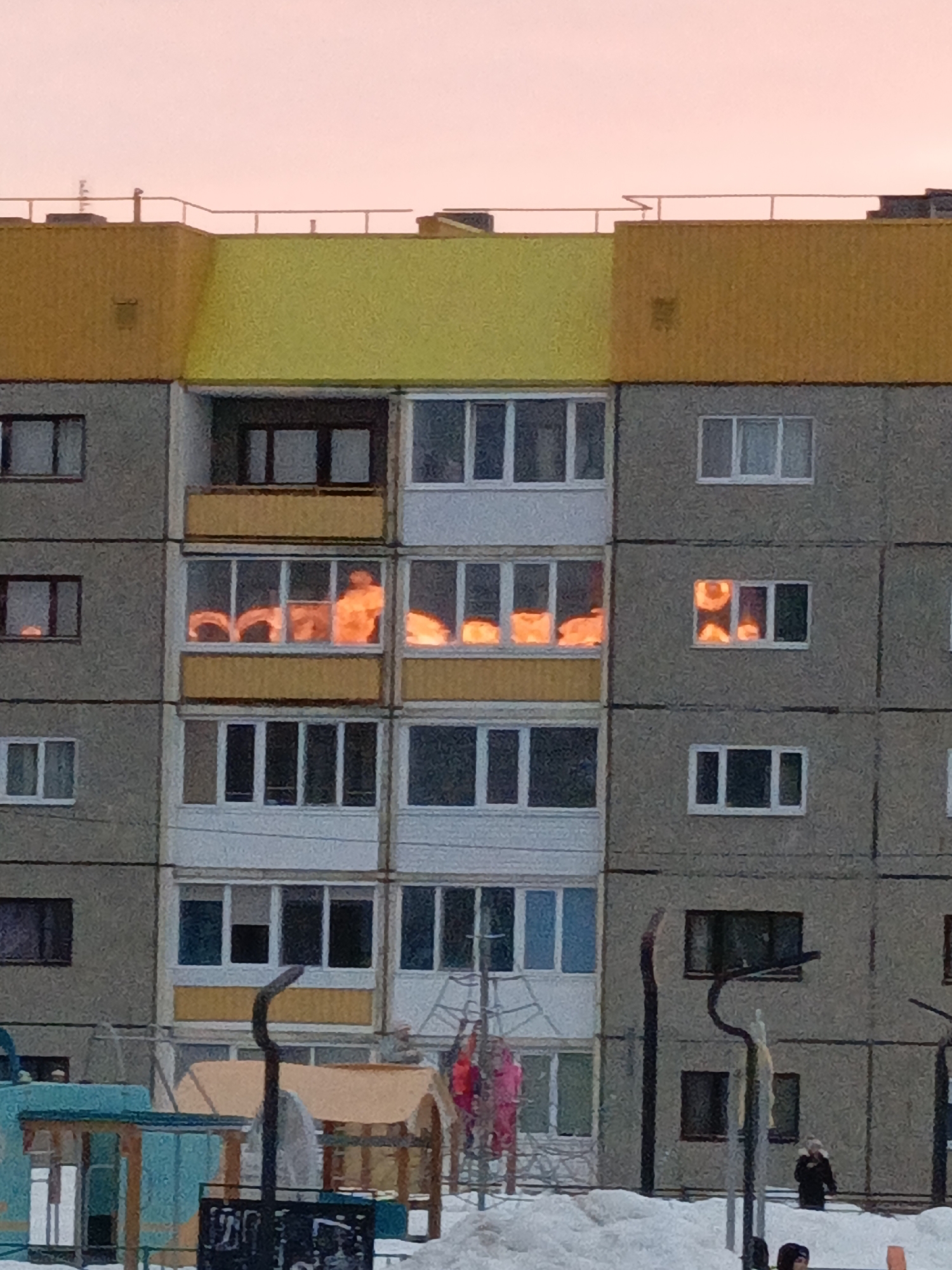 An exterior forensic analysis of an image taken in Марковский, Russia, depicts a multi-story residential building under a pinkish-orange sky, characteristic of twilight. The building's facade is constructed from grey concrete panels, with the top section featuring alternating yellow and orange painted sections and a simple metal railing along the roofline.
Prominently, several windows across multiple floors exhibit intense orange reflections, consistent with either a low-angle sun (sunset/sunrise) or significant internal light sources. Specifically, the second floor from the top shows a balcony with a yellow base, and the two windows to its right display these bright orange reflections. Further to the right, on the same floor, two distinct windows also show similar intense orange light. Below this, the windows of an enclosed balcony on the first floor from the bottom also reflect this strong orange light.
The ground in the foreground is covered in snow, indicating winter conditions. Visible children's playground equipment includes a turquoise-colored structure on the left, a wooden structure, and a metal climbing frame with pink and red elements (possibly garments hanging or part of the equipment). In the lower right corner, two individuals are partially visible amidst the snow and foreground objects; one appears to be an adult in dark clothing, while a smaller figure is partially obscured.