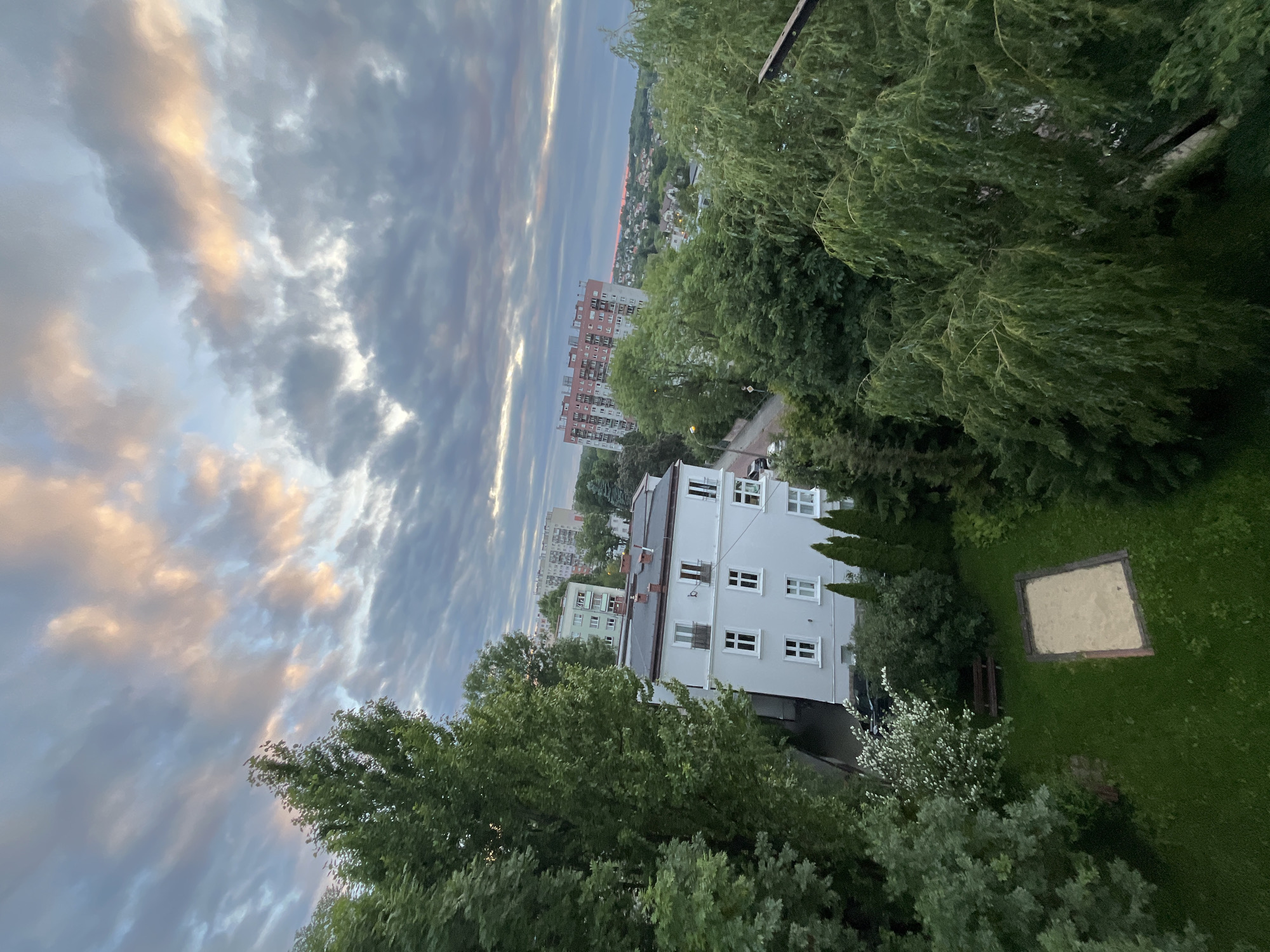 An eye-level, vertical medium shot captures an urban landscape dominated by dense foliage and a dramatic sky at dusk. In the foreground, lush green trees with full canopies frame the scene on the left and right, with a prominent weeping willow on the right. Below, a patch of manicured green lawn features a sandbox with a wooden border and a single wooden bench. A white, multi-story building with multiple windows and balconies is situated behind the lawn. Further in the background, a taller, reddish-brown apartment building with many windows stands amidst a scattering of smaller buildings and more trees, indicating a residential area. The sky above is filled with a complex pattern of clouds, some dark and textured, others illuminated with soft hues of pink and orange, suggesting the last light of the setting sun. The overall impression is one of a serene, yet busy, urban environment at the close of day.