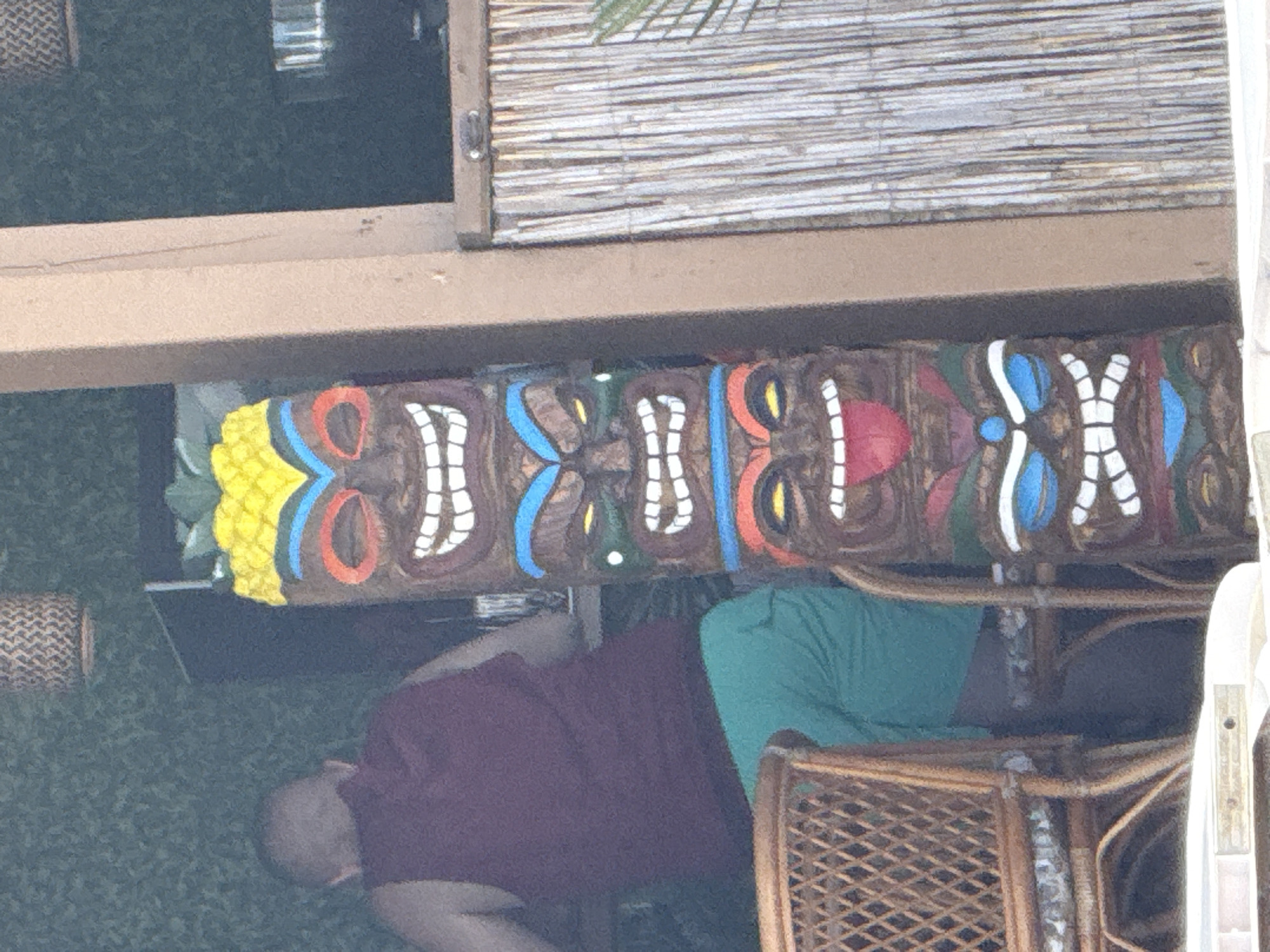 The image shows a close-up, rotated view of a tropical-themed decoration, likely a tiki bar or a similar establishment, in Sozopol, Bulgaria. The primary focus is a row of colorful, carved wooden tiki masks, each with distinct facial features like wide smiles and expressive eyes, painted in shades of brown, red, blue, yellow, and white. One of the tiki masks has a yellow, pineapple-like headdress.
Below the tiki masks, parts of two people are visible, sitting or lounging. One person is wearing a deep red or maroon shirt and has their head down, possibly resting or sleeping. Their legs are partially visible, and they appear to be wearing gray pants. The second person, to the right, is wearing a bright teal or green shirt and is seated in a rattan chair. The chair itself is also made of woven rattan, a common material in tropical decor.
The setting appears to be outdoors, possibly a patio or covered area. In the background, a bamboo-covered wall or screen is visible, further reinforcing the tropical theme. The lighting suggests it could be daytime, though the exact time is not clear. The overall impression is one of a relaxed, vacation-like atmosphere. There is no visible text in the image.