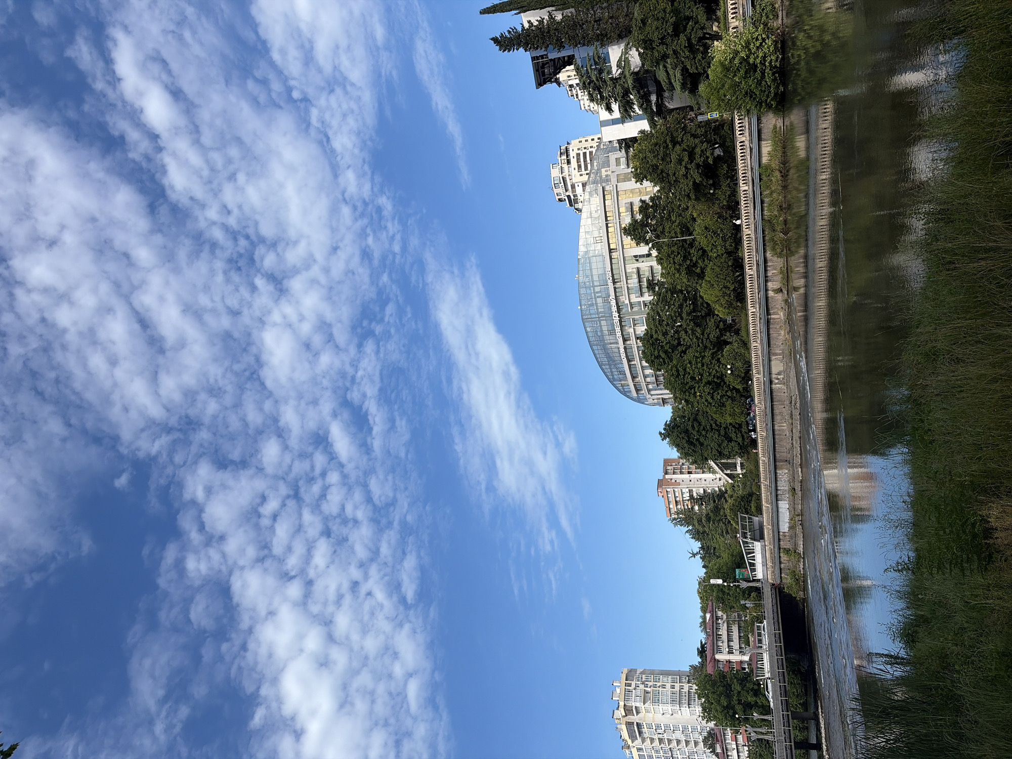This is a vertical, eye-level, full shot of a riverfront cityscape under a blue sky with scattered white clouds. In the foreground, tall green grass lines the riverbank. The river flows with moderate ripples and reflects the surrounding environment. A concrete embankment with an ornate railing separates the river from the city.
A bridge, supported by concrete pillars, spans the river in the mid-ground, with vehicles visible on it. Beyond the bridge, a row of buildings and trees forms the city skyline. The most prominent building is a modern, curved structure with a glass facade and a gold-colored awning. To its left, a tall, light-colored residential building with numerous balconies stands out. Other buildings of varying heights and architectural styles are visible, interspersed with lush green trees. The scene appears to be taken during the daytime on a clear day.
This scene is located in the city of Sochi, Russia, along the river embankment in the central part of the city, likely near the central bridge or the area around the "Zvezdny" hotel, given the architecture of the prominent curved building.