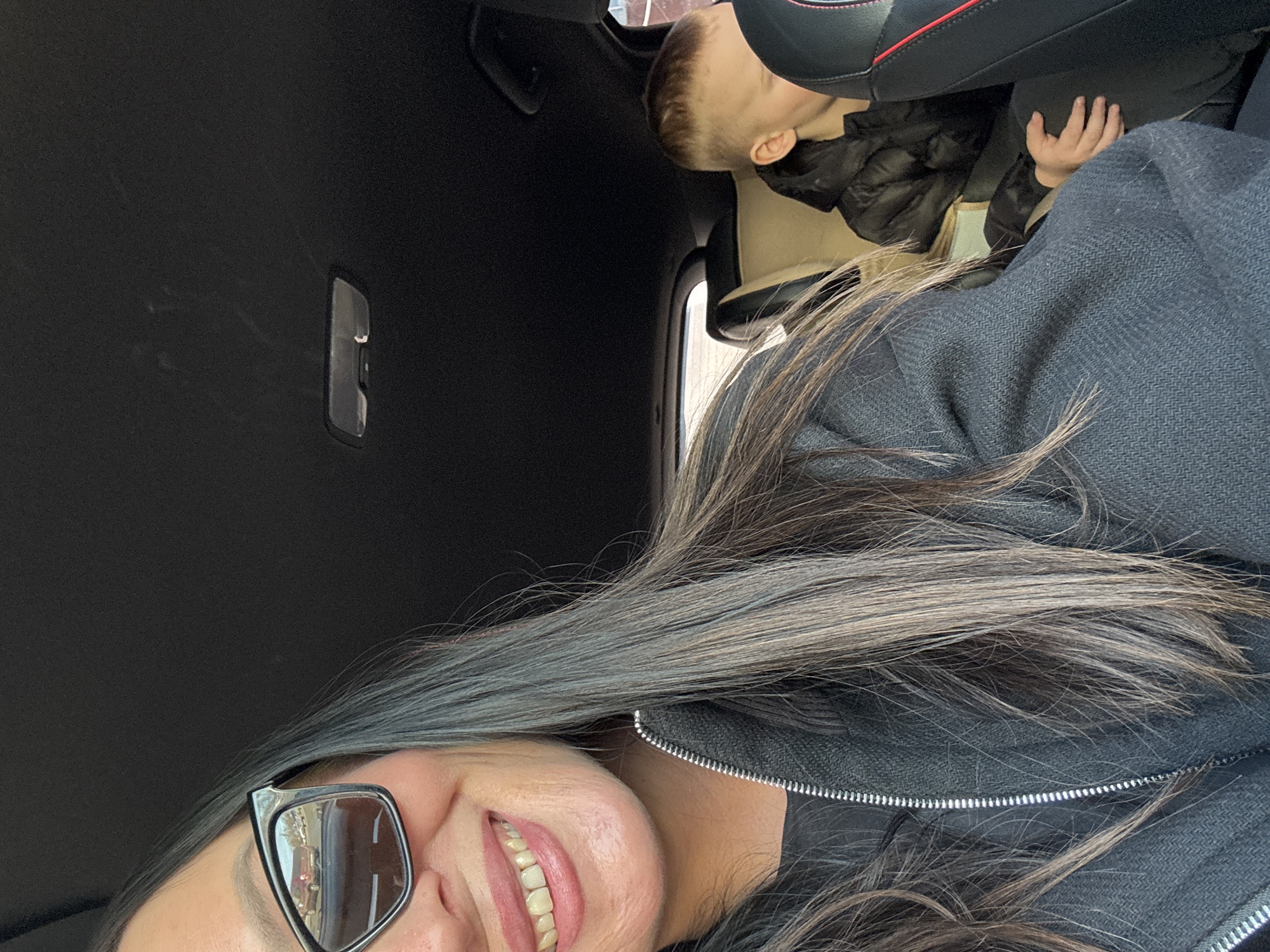 An indoor photograph taken within a vehicle, possibly located in Votkinsk, Russia, captures two individuals. In the foreground, partially cropped on the left, an adult female with long, dark hair and lighter highlights wears black cat-eye sunglasses and a dark gray zippered jacket over a black undershirt. She is smiling, exposing her teeth. The reflection in her right sunglass lens reveals an outdoor scene featuring a red pickup truck, other indistinct vehicles, and distant foliage under a bright sky. In the mid-ground, a child with short, light-colored hair is seated in a rear beige car seat, wearing a black puffer jacket. The child's face is turned downwards, with a right hand resting on their leg. The vehicle's interior includes a black headliner and an overhead light fixture. A black seat with red stitching is partially visible on the extreme right.