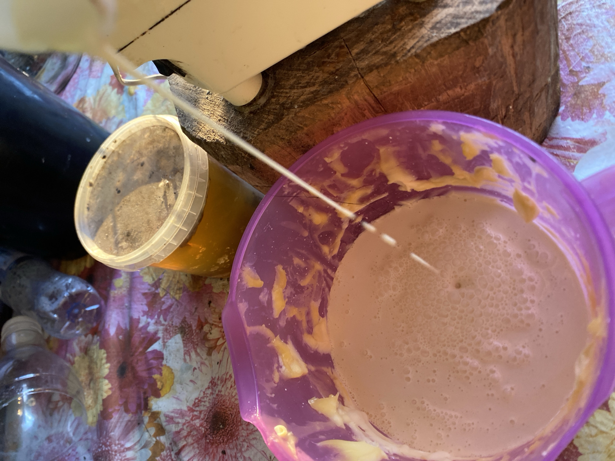 The image shows a close-up of a purple bowl filled with a frothy, light-colored liquid, possibly a batter or smoothie. Bits of yellow material are adhered to the inside of the bowl. A thin stream of liquid is pouring into the bowl from an unseen source, likely from a blender or dispenser, which is partially visible on the right side of the frame. Behind the bowl, there is a clear plastic container filled with amber-colored liquid, possibly honey, with a slightly dusty lid. The objects are situated on a surface covered with a floral-patterned tablecloth. In the background, parts of plastic bottles and a dark container are visible. The overall scene suggests a food preparation or processing activity. The location of Maloarslangulovo, Russia, is not evident from the image itself.