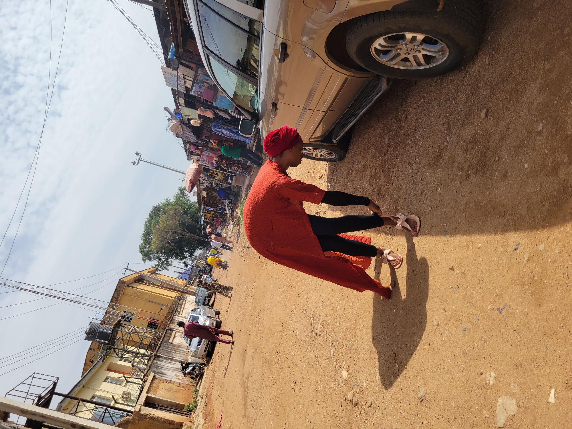 The image shows a view of a street scene in Jos, Nigeria. A woman wearing a red head covering, an orange long-sleeved tunic, and black pants is seated on the ground next to a parked tan SUV. She is wearing sandals and appears to be adjusting them. The ground is a sandy-brown color, consistent with dirt or unpaved terrain.
In the background, there's a crowded marketplace visible, with numerous people and umbrellas. Buildings are visible in the distance. A light pole is to the left. Other cars and individuals walking are present in the middle distance. The sky is partly cloudy.