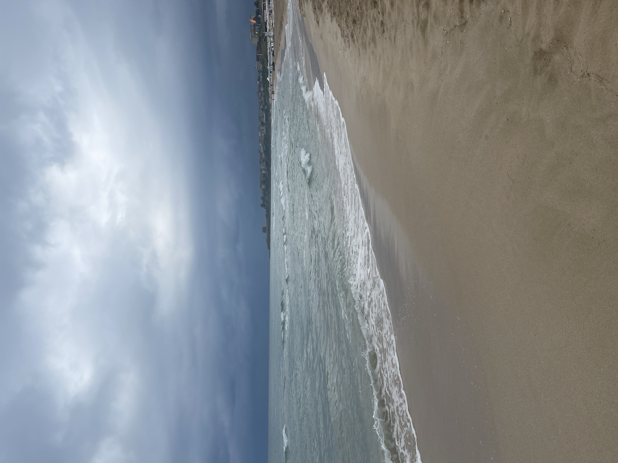 The image depicts a beach scene in Primorsko, Bulgaria, under a heavily overcast sky. The foreground is dominated by a sandy beach, wet near the water's edge, with fine footprints visible on the dry sand further back. Gentle waves with white foam are breaking on the shore. The sea is a muted grey-blue, with whitecaps suggesting a moderate breeze. In the distance, along the coastline, a town or resort area is visible, characterized by buildings with red-tiled roofs and a scattering of white umbrellas and lounge chairs on a more populated section of the beach. Several indistinct figures can be seen on the distant beach. A prominent tower-like structure, possibly historical or a landmark, stands on a rocky outcrop to the left of the town. The sky is filled with dark, heavy clouds, indicating potential rain or stormy weather. The overall mood is somber and atmospheric, typical of a cloudy day at the seaside.