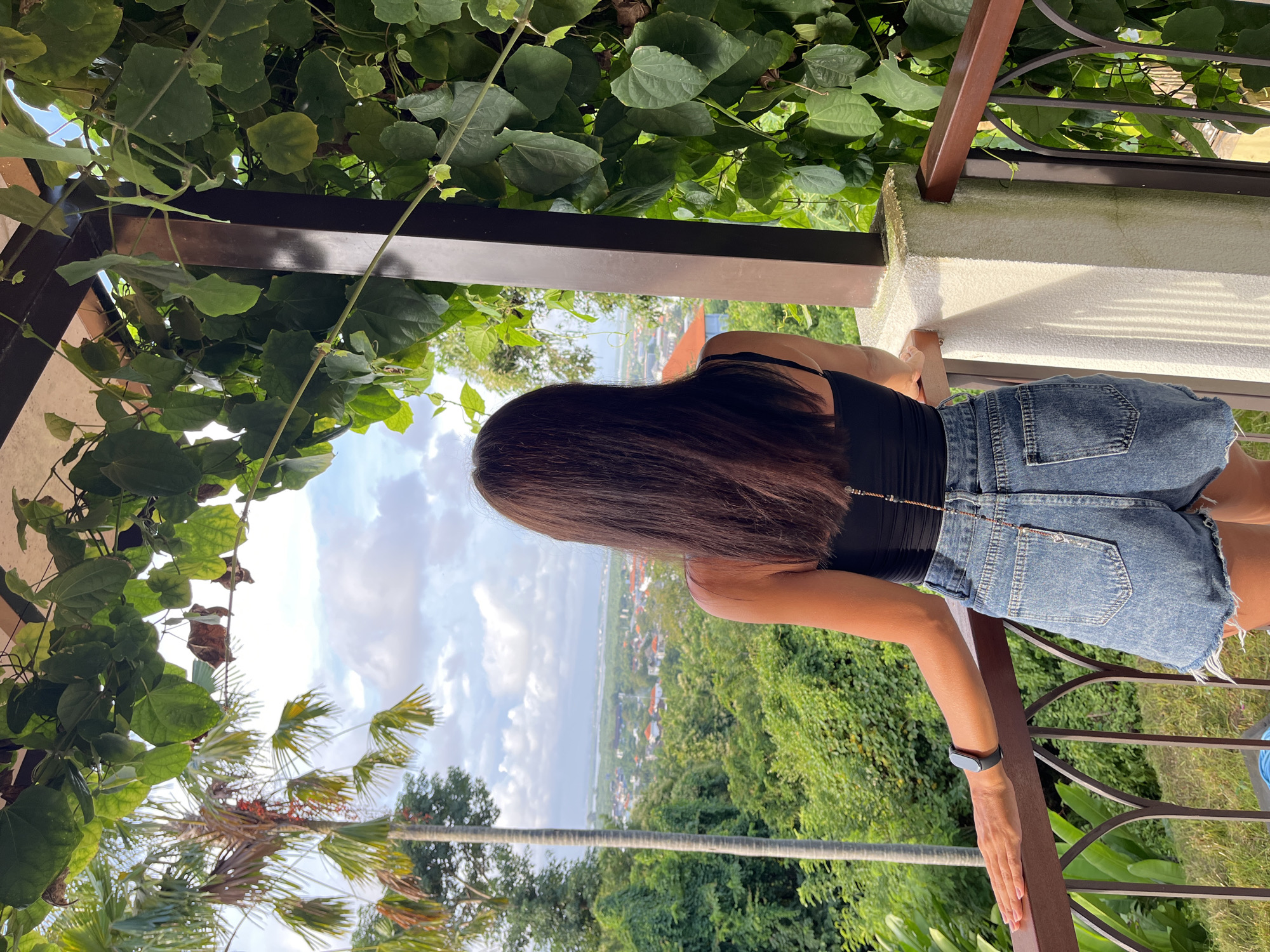 A woman stands on a balcony overlooking a landscape in Kuta Selatan, Indonesia. The woman is seen from the back, with long brown hair. She is wearing a black tank top and denim shorts, leaning against a white wall and metal railing. A fitness tracker is visible on her wrist. The foreground features lush green foliage, with vines and palm trees framing the scene. The background showcases a cityscape blending into a green hillside and the ocean under a partially cloudy sky. A brown metal structure, possibly part of the balcony, is visible overhead.