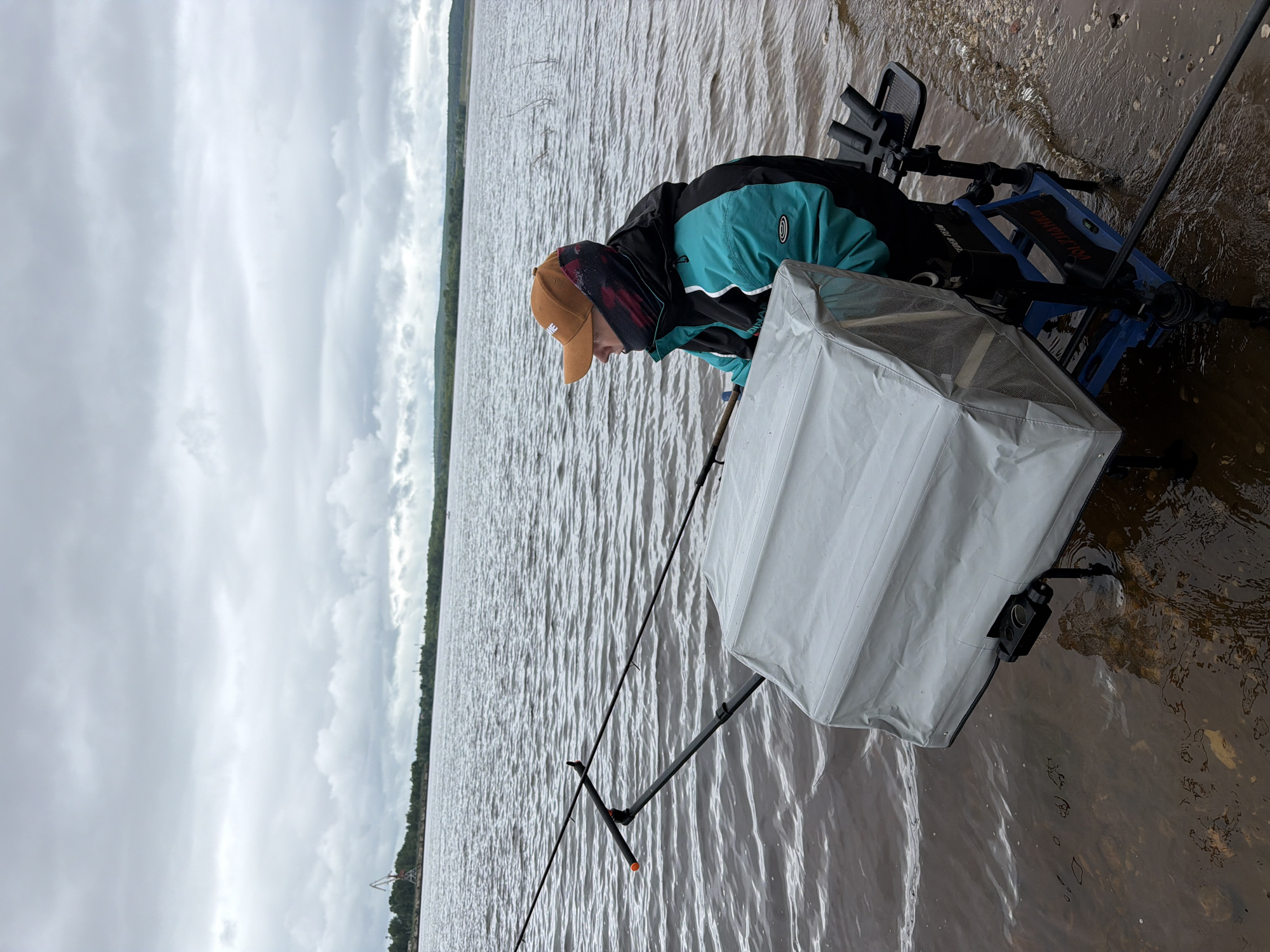 An individual is depicted fishing from the bank of a wide river under a uniformly overcast sky. The person, seen in right profile, wears a teal and black jacket, a brown baseball cap, and a dark patterned neck gaiter covering their lower face. They are seated on a blue and black multi-legged fishing platform, or 'feeder chair', with its legs partially submerged in the shallow, dark brown river water. Two fishing rods extend from the platform over the water, supported by an attached rod rest system. A large, rectangular, light grey fabric-covered container, likely a bait box or side tray, is affixed to the left side of the platform. A smaller black accessory tray with multiple rod holders is attached to the right. The immediate shoreline is composed of dark, wet soil and pebbles. Across the river, a distant treeline forms the horizon. A prominent red and white gantry crane is visible on the far left of the distant bank, indicating an industrial port area. Further along the distant bank, additional industrial structures are faintly visible. The sky is completely covered by a dense layer of grey clouds. This scene is located on the Kama River, facing the industrial port area, in the city of Chaikovsky, Russia.