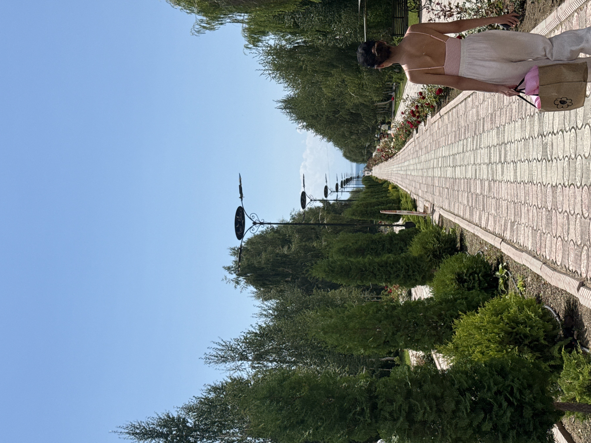 A young woman walks away from the camera down a paved walkway lined with trees and rose bushes. She is wearing a pink camisole and loose white pants, and carrying a large brown tote bag. The walkway extends into the distance, where it appears to lead towards a body of water. The scene suggests a park or garden setting in Sary-Oy, Kyrgyzstan, characterized by its lush greenery and clear blue sky.
