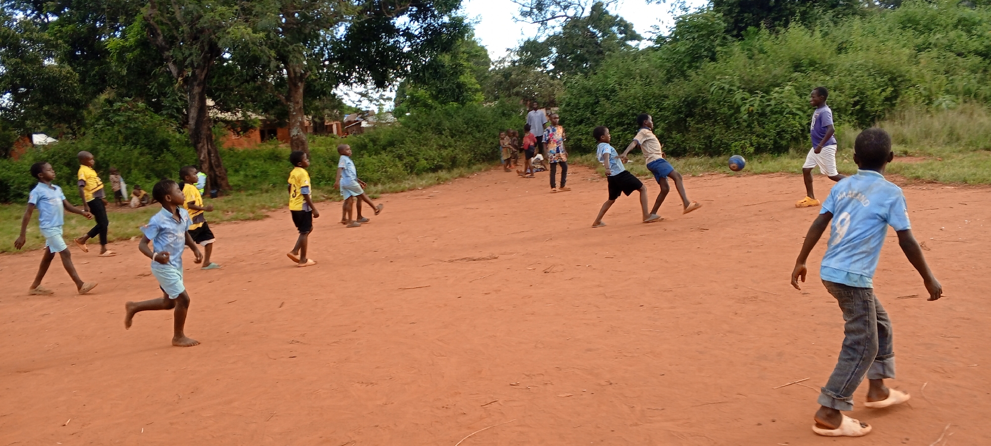 In Ngalula, Democratic Republic of the Congo, a group of children is playing soccer on a dirt field. The field appears to be an open area adjacent to some trees and brush. The children are dressed in various clothing, including blue and yellow shirts, shorts, and jeans. Some children are barefoot while others wear sandals. One child is kicking a soccer ball. In the background, there is a group of adults and children watching the game.