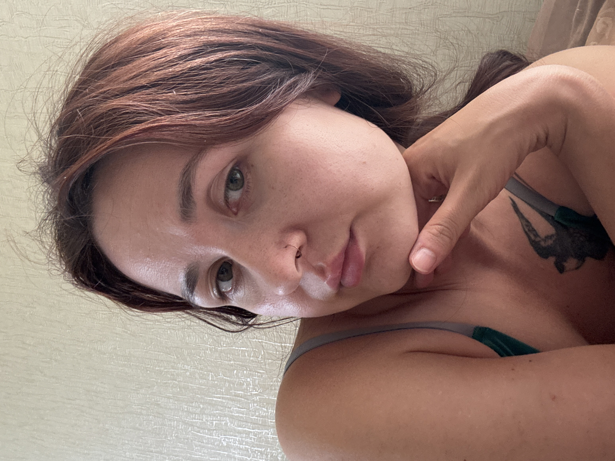 This is a close-up selfie of a woman with brown hair and green eyes. She is lying down and resting her chin on her hand. She is wearing a dark green top with thin straps. A tattoo of a bird is visible on her chest. The background appears to be a textured wall. The scene is indoors, likely in a bedroom, and there are no visible indicators of the city of Naberezhnye Chelny, Russia.