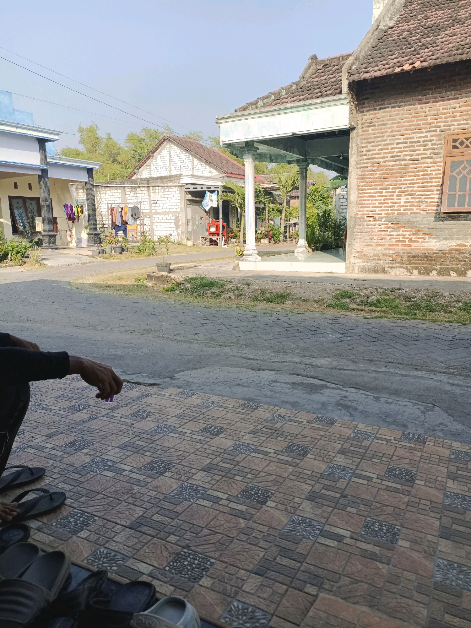 The image depicts an outdoor scene on a sunny day, likely in Rancang Kencono, Indonesia. In the foreground, a person, partially visible and dressed in dark clothing, is sitting on a tiled patio. They are holding a small purple object in their right hand. A collection of footwear, including flip-flops and sandals, is arranged on the tiles.
The middle ground shows a paved pathway or road. Beyond the path, several houses are visible. To the left, a light-colored house with a blue roof trim has clothes drying on a line. Adjacent to it, a house with white brick walls also features clothes hanging on a rack. Further back, greenery, including trees and plants, is scattered throughout the scene. A prominent structure to the right is a house constructed with red bricks, featuring a traditional tiled roof and a wooden-framed window. A covered porch area with pillars is visible in front of this brick house. The sky is clear and blue.