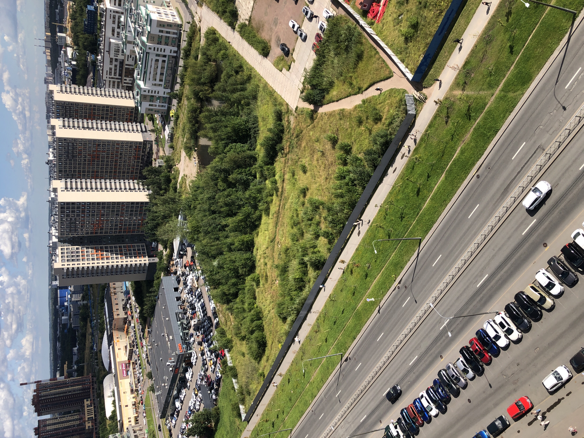 This is an aerial view of an urban environment, likely a suburban area, with a focus on a multi-lane road and adjacent green spaces and buildings. The road, running diagonally across the lower half of the image, has several lanes marked with white lines and a metallic fence bordering its grassy median. A line of parked cars is visible along the right side of the road.
Beyond the road, a wide expanse of grass and trees stretches towards a cluster of modern, high-rise residential buildings. These buildings are tall, with numerous windows and a mix of beige and grey facades, some featuring orange accents. One building appears to have a distinctive chimney-like structure behind it. In the middle ground, a car dealership with a grey building and many vehicles parked in front is visible, along with a large retail complex with a white arched roof. Further in the distance, more buildings and industrial structures can be seen, suggesting a developed urban landscape. The sky is partly cloudy with scattered white clouds against a blue background.
Several people are visible walking on pathways adjacent to the road and near the buildings. A few cars are also seen driving on the road and parked in various lots. The overall scene depicts a contemporary urban development with a clear separation between infrastructure, green areas, and residential and commercial structures.