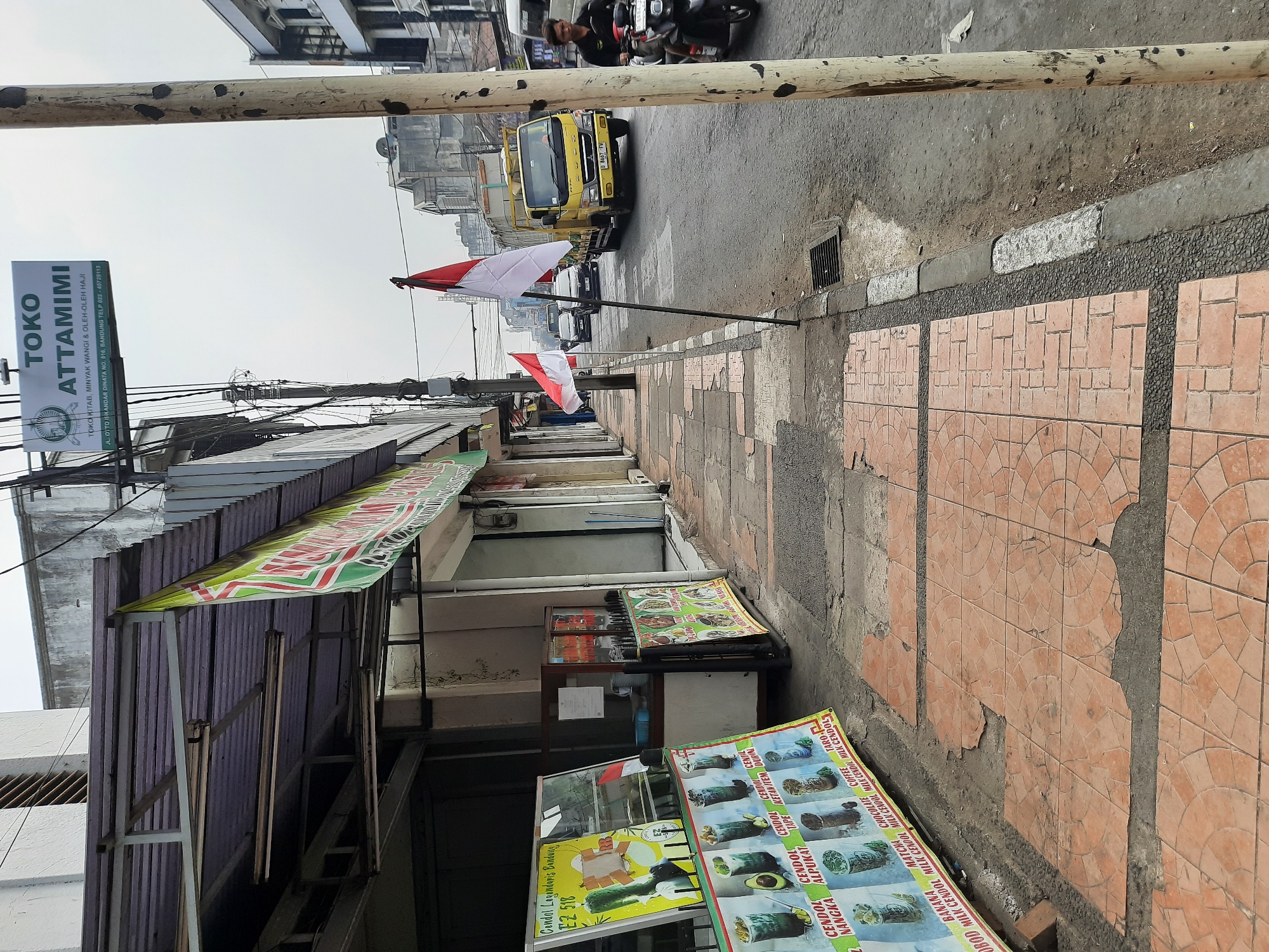 The image depicts a street scene in Bandung City, Indonesia. In the foreground, a sidewalk with broken tiles is visible, leading towards a row of shops. One shop has a banner displaying images of various types of "Cendol," a traditional Indonesian dessert. Further down the sidewalk, a sign for "TOKO ATTAMIMI" is prominently displayed, indicating a store selling books, perfumes, and gifts for Hajj pilgrims.
The street is paved and has a drainage grate. Indonesian flags are planted along the sidewalk, suggesting a national celebration or event. A yellow truck is driving on the street, followed by other vehicles. A person on a motorcycle is also visible, heading towards the viewer. The buildings lining the street appear to be commercial establishments, with some showing signs of age and wear. The sky is overcast, indicating a cloudy day.