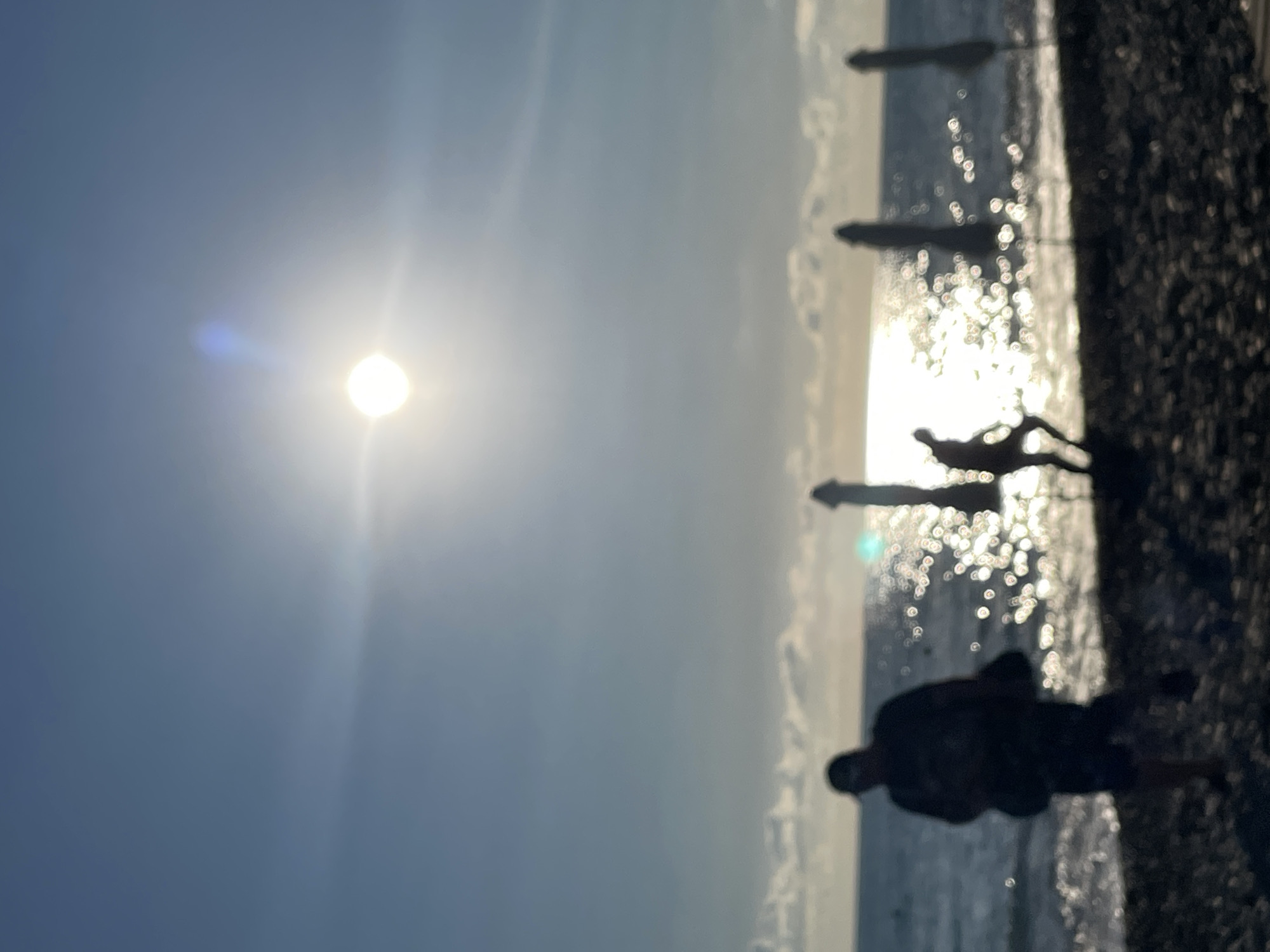 The image depicts a beach scene with the sun high in the sky, casting a bright glare on the water. Silhouetted figures of people and beach umbrellas are visible along the shoreline. The foreground consists of pebbles on the beach, while the middle ground shows the gentle waves of the ocean reflecting the sunlight. In the background, the horizon separates the sea from the sky, which is a clear blue with a few wispy clouds. The overall impression is one of a sunny day at the beach.