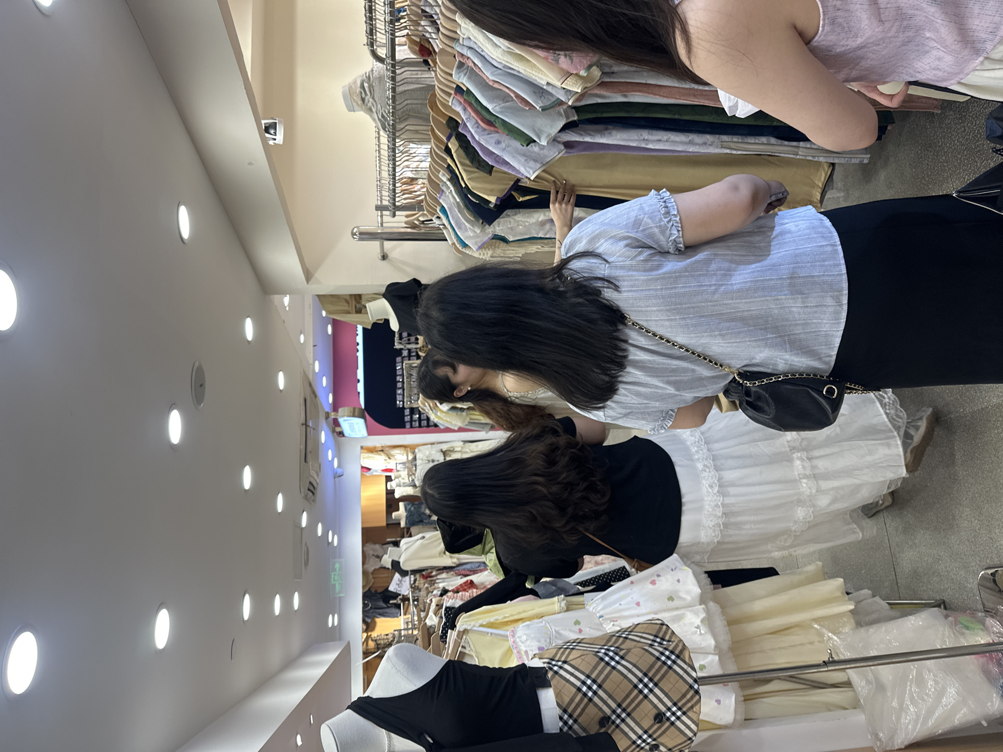 In a brightly lit clothing store located in the Chongqing High-tech Zone, China, multiple people are browsing through racks of clothes. One woman, wearing a light blue striped shirt and a black skirt, is reaching for a garment on a rack filled with various colored shirts. Her black purse hangs at her side. Another woman, wearing a white top, stands next to her, also looking at the clothing rack. A third woman, dressed in a black top and white lace skirt, is looking at clothing on a lower rack. In the foreground, there is a mannequin dressed in a black top and a plaid skirt. The store has bright recessed lighting.