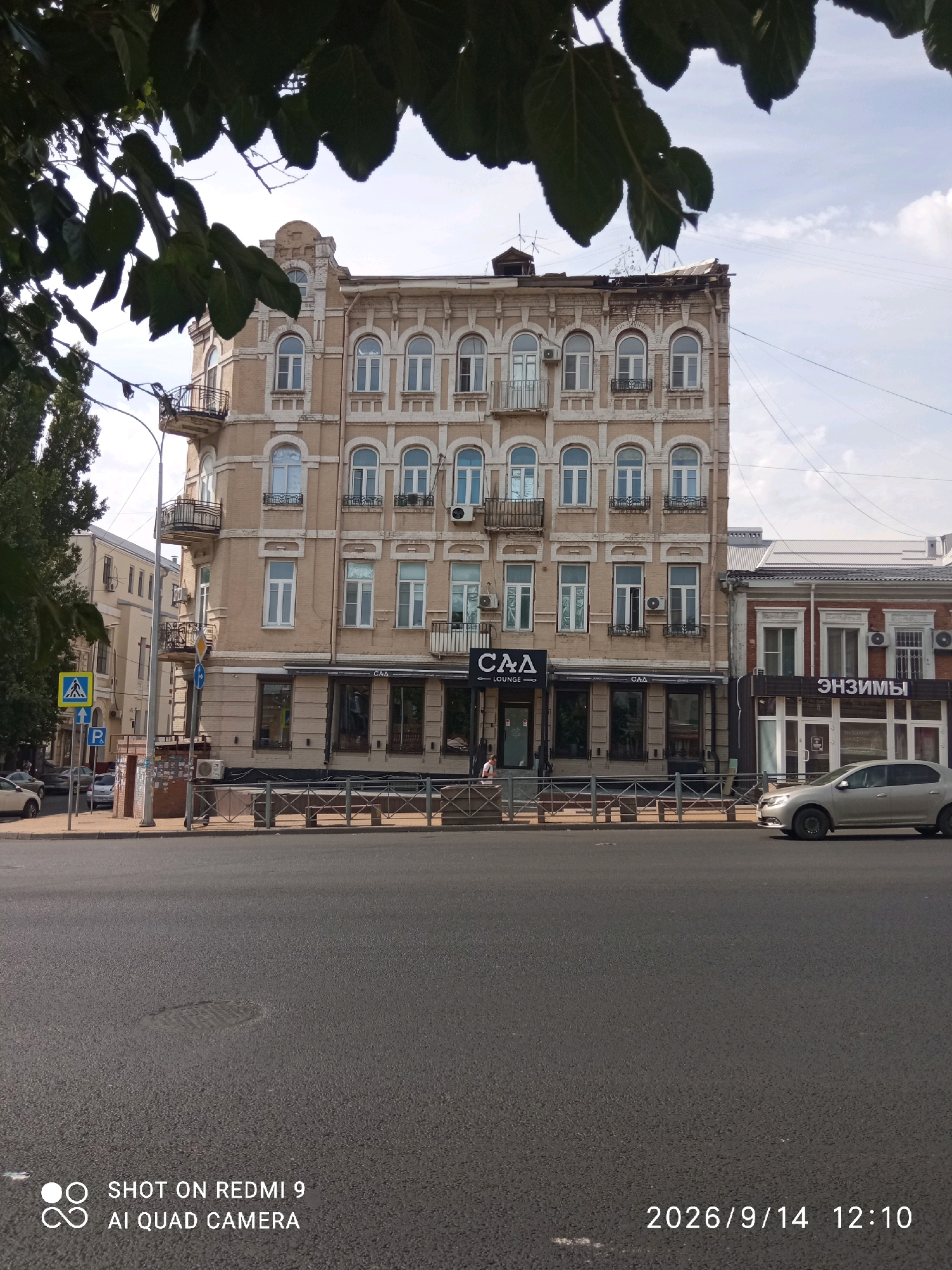 The image captures a street scene in Rostov-on-Don, Russia. In the foreground, a dark asphalt road dominates the lower half of the frame, with a manhole cover visible. The middle ground features a sidewalk with a decorative metal fence. Behind the fence, a tan-colored, multi-story building with arched windows and balconies is prominent. A sign reading "САД LOUNGE" is affixed to the building's facade. To the right of this building, a red-brick building with large windows and the Russian word "ЭНЗИМЫ" (Enzymes) on its facade is visible. A silver sedan is parked in front of the red-brick building, and another car is partially visible on the left side of the street. In the background, a taller, light-colored building with multiple windows and balconies is seen. Trees with large green leaves partially obscure the top of the image. The sky above is a pale blue with scattered white clouds. A pedestrian crossing sign and a blue sign with a white 'P' are visible on the left side of the street. The overall impression is of an urban street on a bright, possibly warm day.
