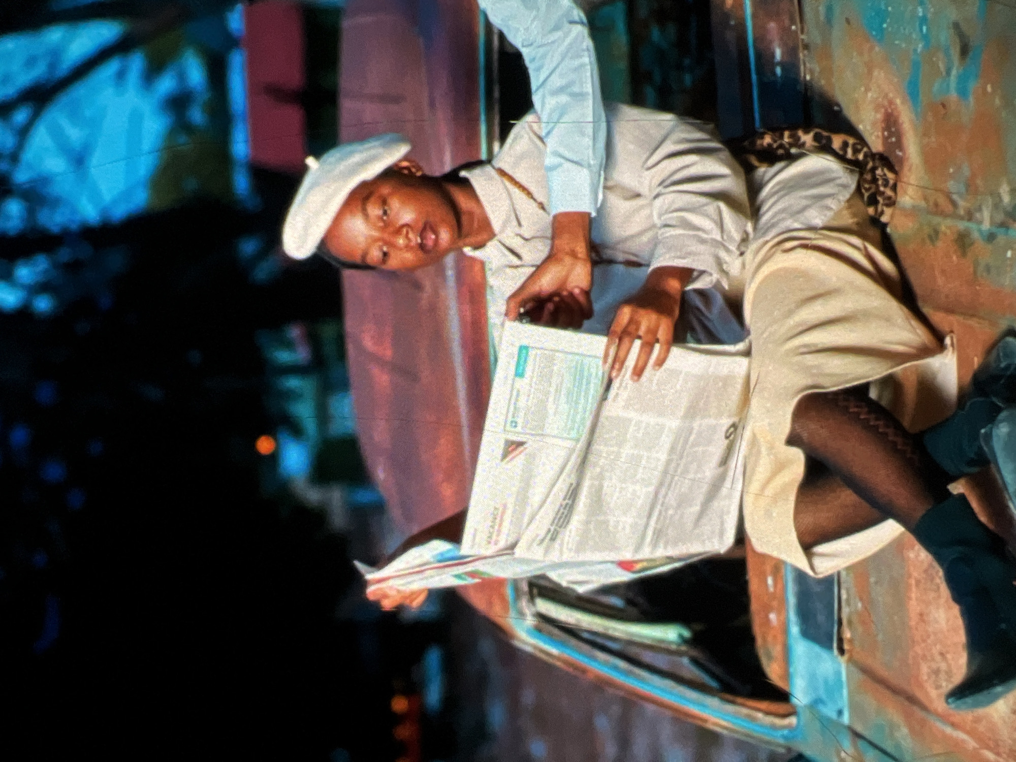 A young Black woman wearing a white beret and a light-colored button-up shirt sits on the hood of a rusty, vintage car. She is holding a newspaper, with one hand pointing to an article and the other supporting the paper. Her legs are crossed, and she is wearing dark, patterned stockings and black boots. A second person's arm, wearing a light blue shirt, is visible reaching towards the newspaper. The car appears to be in an outdoor setting, possibly a scrapyard or an abandoned area, with a blurred background of trees and some distant lights, suggesting an urban environment. The lighting is dramatic, with the scene illuminated by artificial light sources, creating strong shadows. The car is old and shows significant signs of rust and decay, with parts of the interior and exterior visible. The overall impression is one of a stylized, artistic composition. The presence of a vintage car and the general urban decay could be indicative of various locations, but without specific landmarks, a precise location within Lagos, Nigeria cannot be determined.