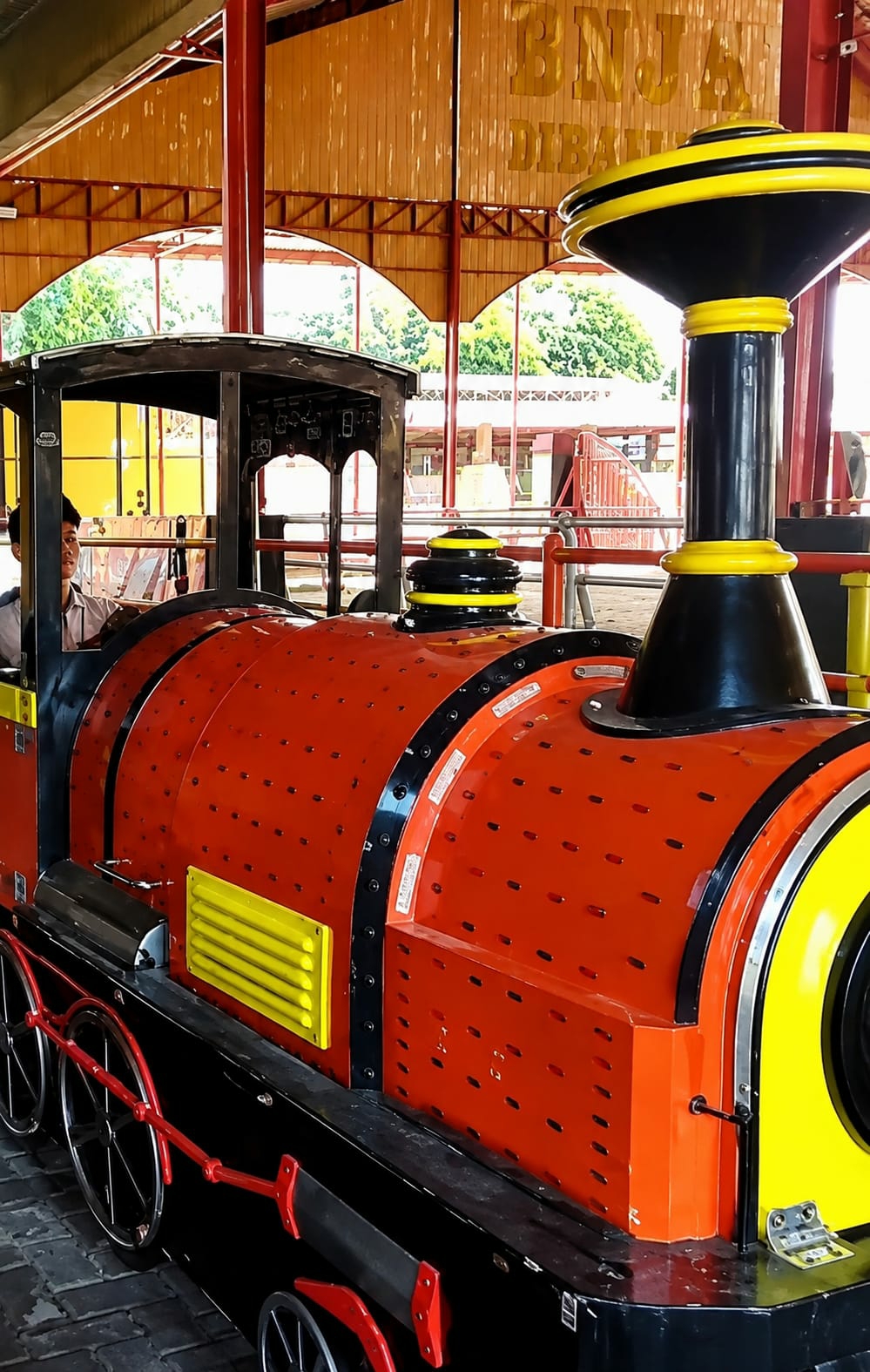 The image shows a close-up of a red and black toy train with yellow accents. The train is parked under a covered structure with a wooden ceiling and metal supports. In the background, there is a sign that reads "BNJA DIBA..." which is likely an abbreviation for "City of Binjai". A person is visible sitting in the driver's cabin of the train. The train appears to be at an amusement park or a tourist attraction in the City of Binjai, Indonesia. The overall impression is that of a fun and family-friendly environment.