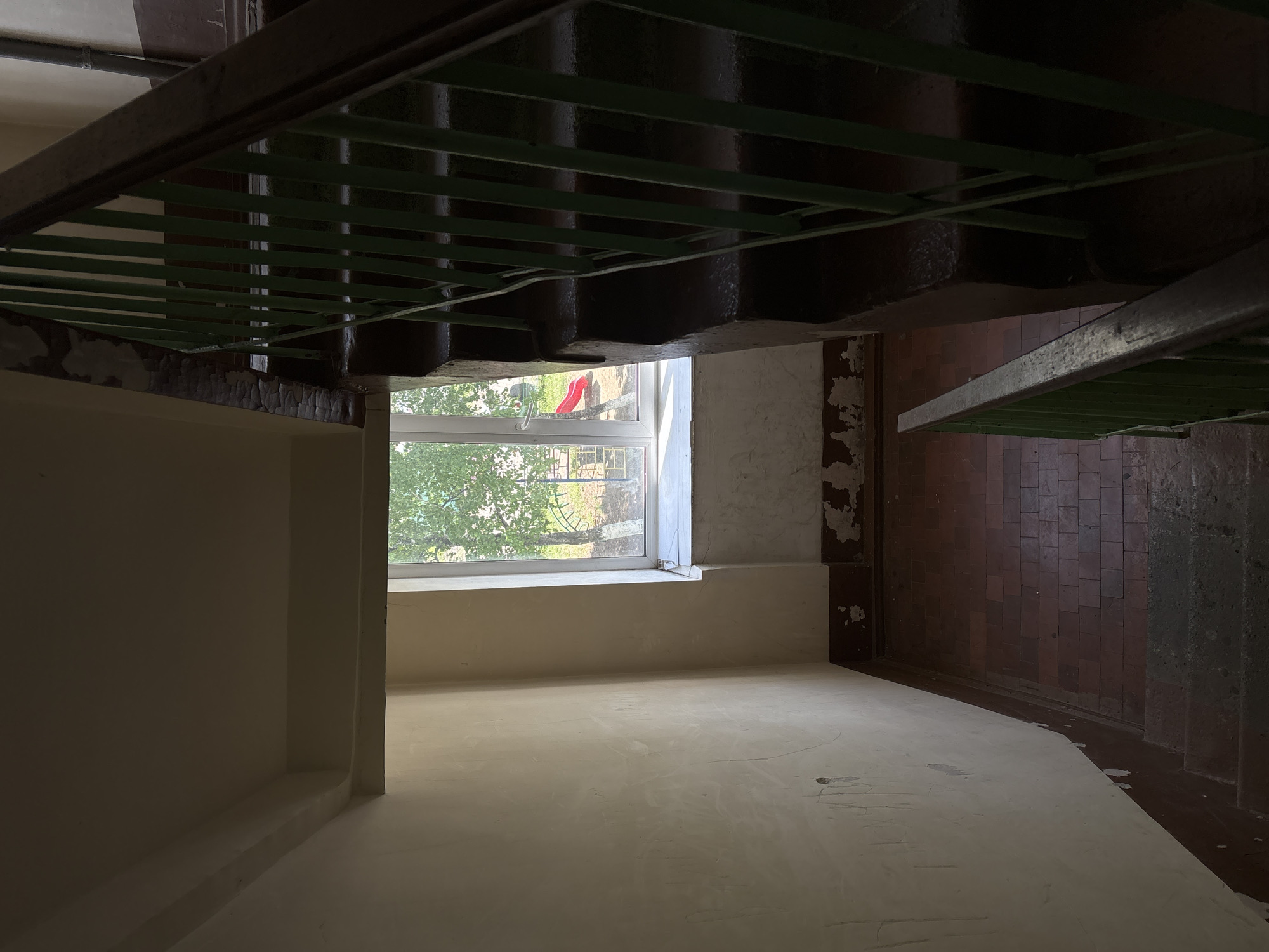 The image depicts the interior of a building, potentially a stairwell or hallway, in Unknown, Unknown. The perspective is looking up from a low angle. A window is visible, with green foliage outside. A red object can be seen through the window. The walls are mostly white, with peeling paint in areas. A staircase is visible, with green metal supports. The floor is mostly off-white. Brown tiles are visible near the staircase.