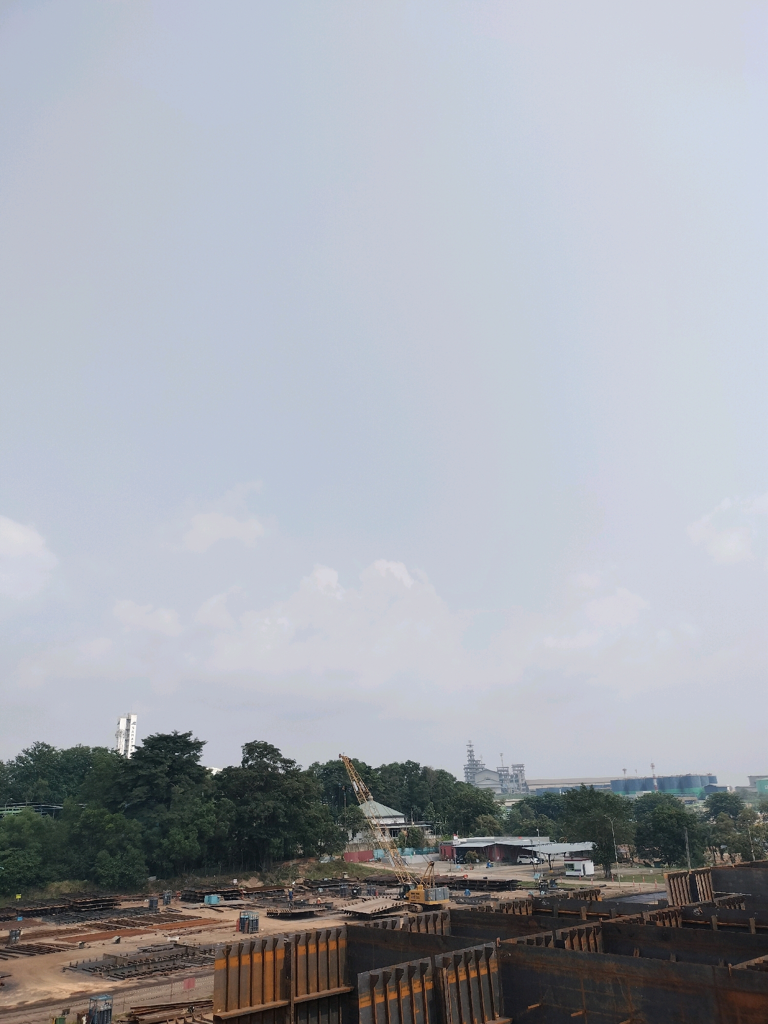 In Batam, Indonesia, an outdoor construction site is depicted with stacks of metal beams and panels laid out on the ground. A yellow crane is visible amidst the greenery of trees in the background. Further in the distance, industrial buildings with tall structures and pipelines can be seen, hinting at an industrial zone. The sky is overcast, with a few scattered clouds. The overall scene suggests ongoing construction or industrial activity.