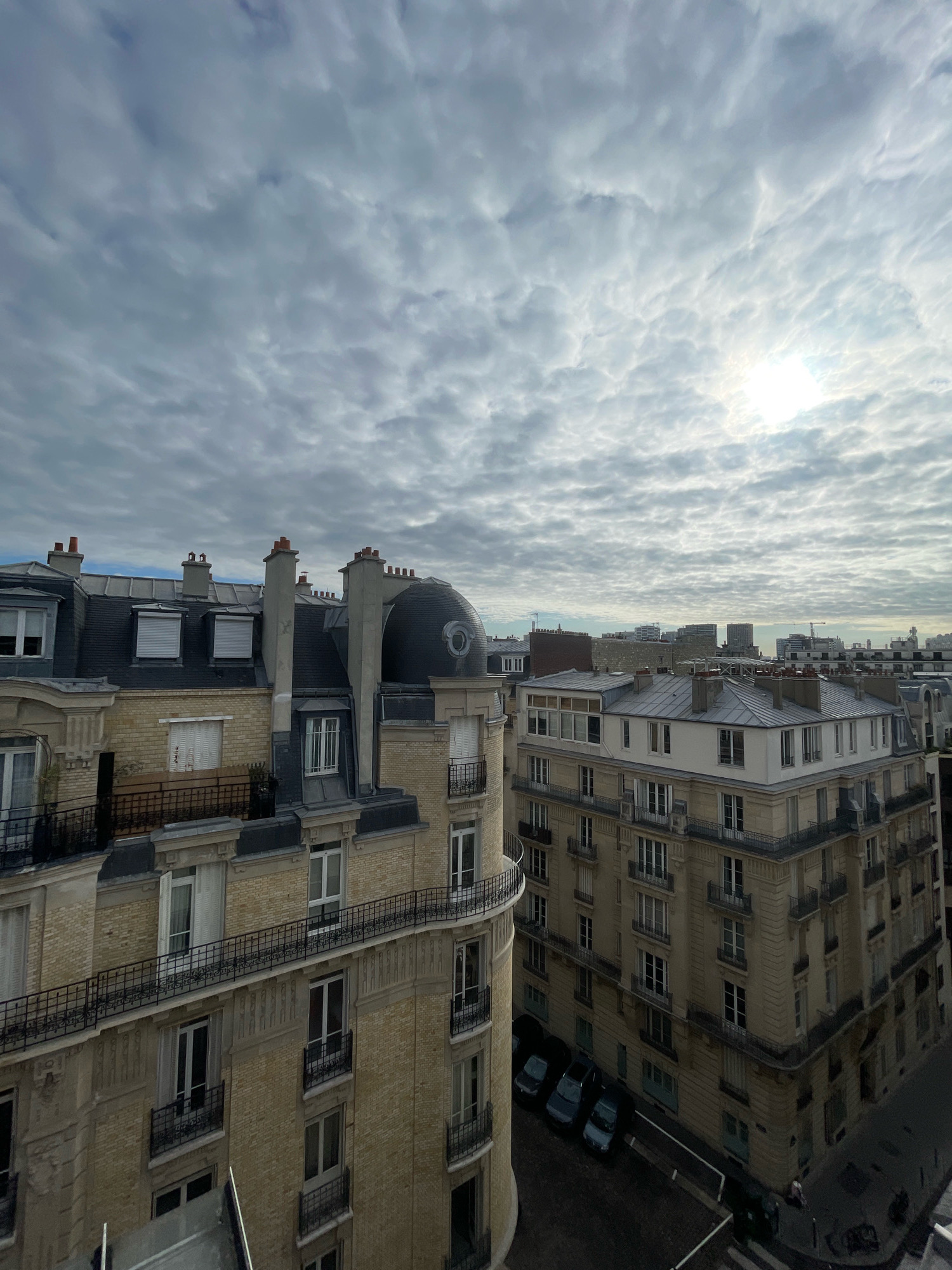 This image depicts a view of Parisian rooftops under a cloudy sky with the sun partially visible through the clouds. The architecture is characteristic of Paris, with mansard roofs, dormer windows, and decorative facades. In the foreground, two buildings are prominent. The building on the left has a light-colored brick facade with several rows of windows and balconies with black metal railings. Chimneys are visible on its roof, along with a distinctive domed structure. The building on the right is taller and also features multiple stories with windows and balconies. Its facade is a light cream color with ornate detailing. A street is visible at the bottom, with several parked cars and a pedestrian. The overall scene suggests an urban environment in Paris, likely residential or commercial, captured from an elevated viewpoint.