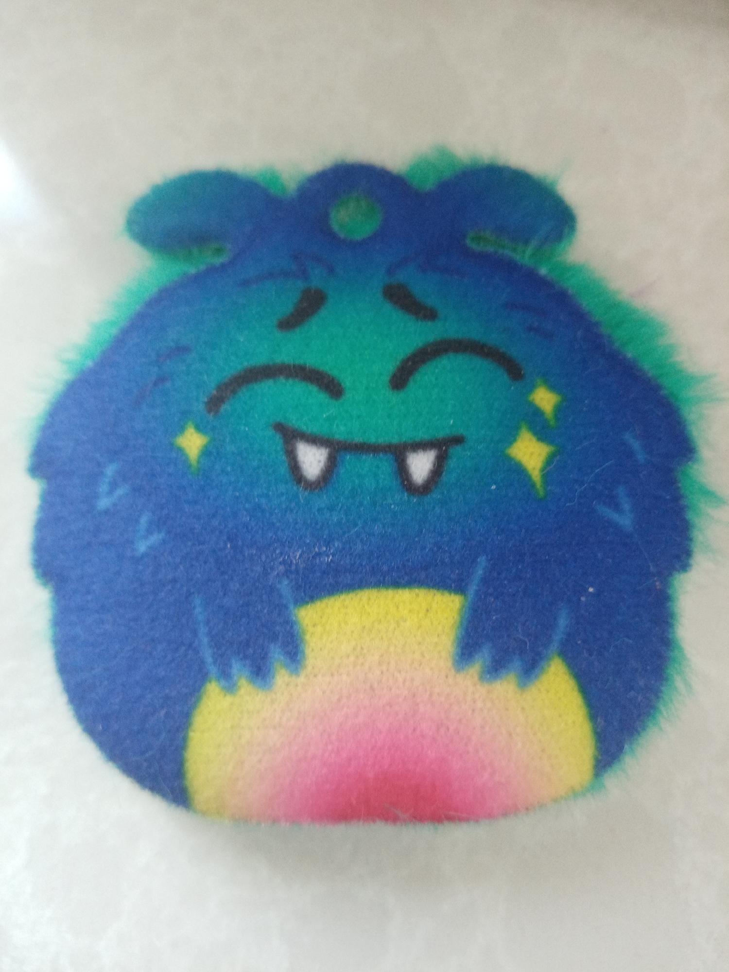 A close-up image shows a stuffed animal toy. It has a blue and green furry exterior, a cartoon face with closed eyes, and small fangs. The bottom of the toy fades from yellow to pink. The toy is resting on a white surface. There is no indication of this image being taken in Elektrostal, Russia.