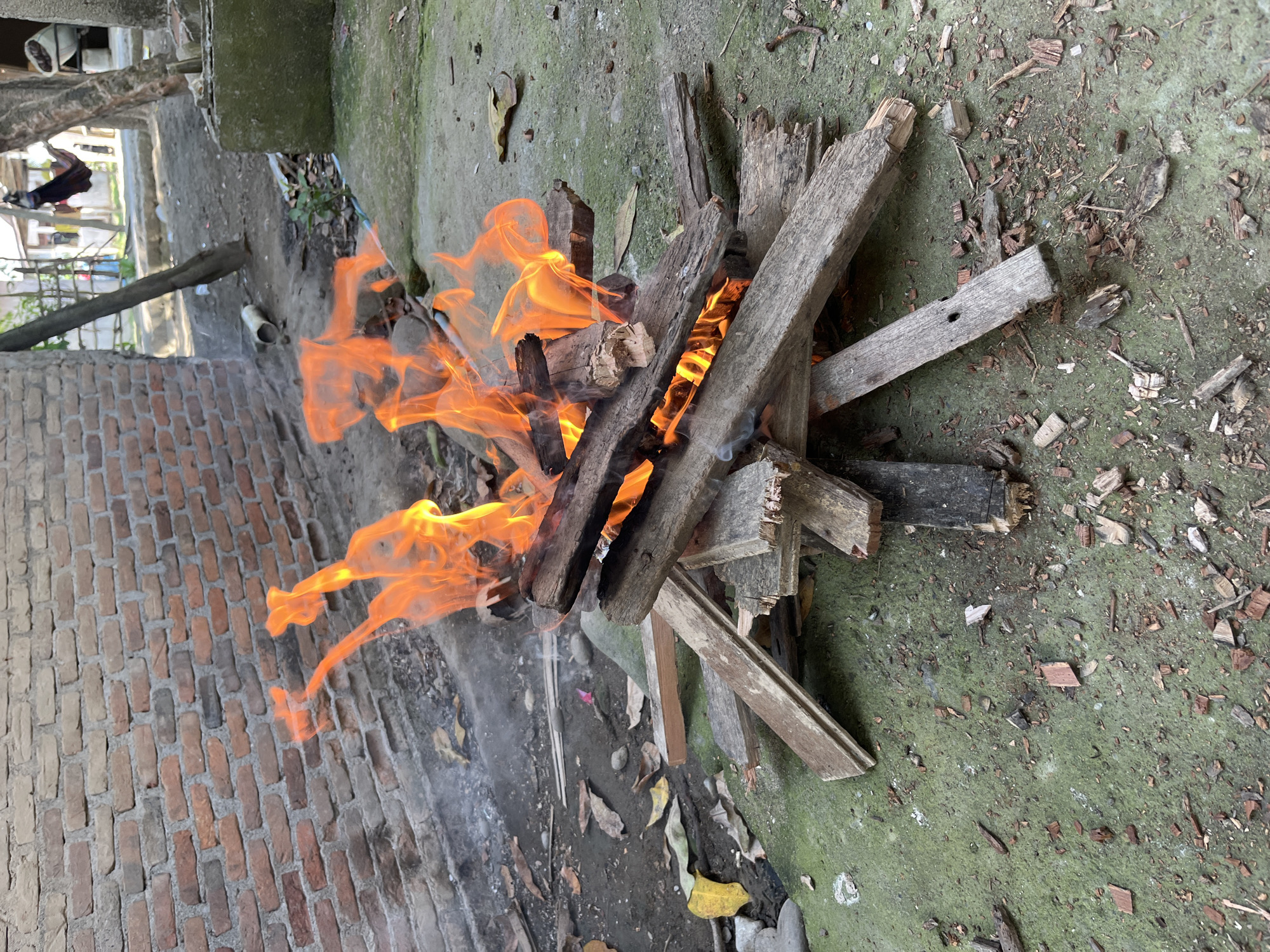 An outdoor scene in Kruengmate, Indonesia, features a small bonfire constructed from discarded wooden planks. The fire is actively burning, with bright orange flames licking upwards. The bonfire is situated on a patch of green, mossy ground, with scattered debris of dried leaves, small stones, and wood shavings. Behind the fire, a weathered brick wall extends horizontally across the frame. To the right, a dirt path is visible, leading towards a rustic, outdoor setting with trees and a partially obscured structure. The overall impression is one of a simple, utilitarian fire in a rural or semi-rural environment.