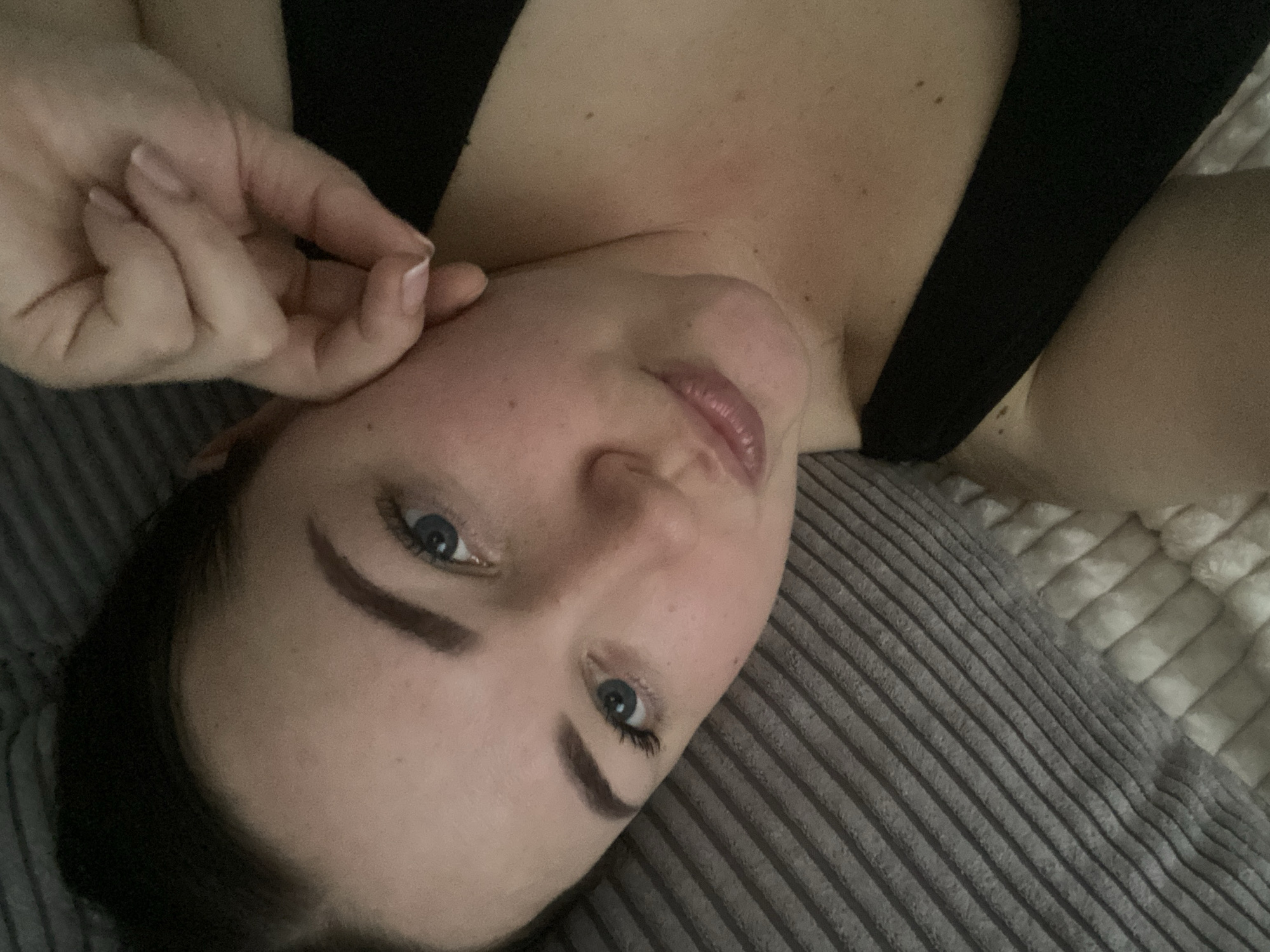A young adult female is depicted in a close-up, angled shot, lying on what appears to be bedding. She has fair skin, dark hair, blue eyes, defined eyebrows, and full lips, which are slightly pursed. She is wearing a black top, with a portion of the strap visible on her right side. Her right hand is raised, with fingers bent, and her thumb is pressed against her left cheek/jawline, causing a slight indentation and localized redness on her skin. She gazes directly forward. The background consists of a grey, horizontally ribbed textile (possibly a pillow or blanket) on the left and a white, fluffy textured fabric in the lower right, suggesting an indoor setting like a bedroom. The specific location within the city of Sochi, Russia, cannot be determined from the visual content of this image.