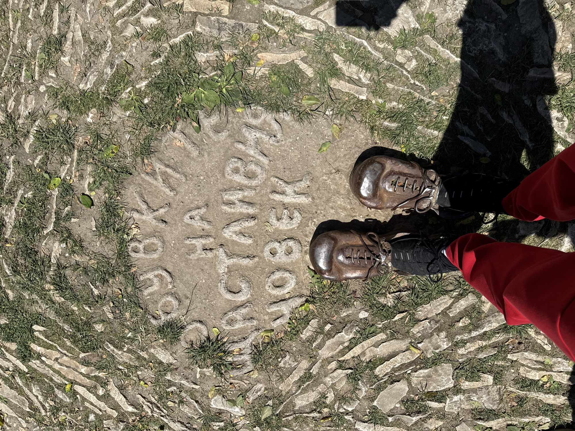 A full shot presents a concrete circle embedded in a cobblestone-paved area, potentially a pedestrian walkway, with a pair of feet standing on either side. The text "по стъпките на П. К. Яворов 1891" is embossed into the concrete circle. The feet belong to a person wearing red pants and black shoes with brown metal-look shoe covers. The shadows cast by the individual and objects suggest outdoor lighting. The location is Lukovit, Bulgaria.