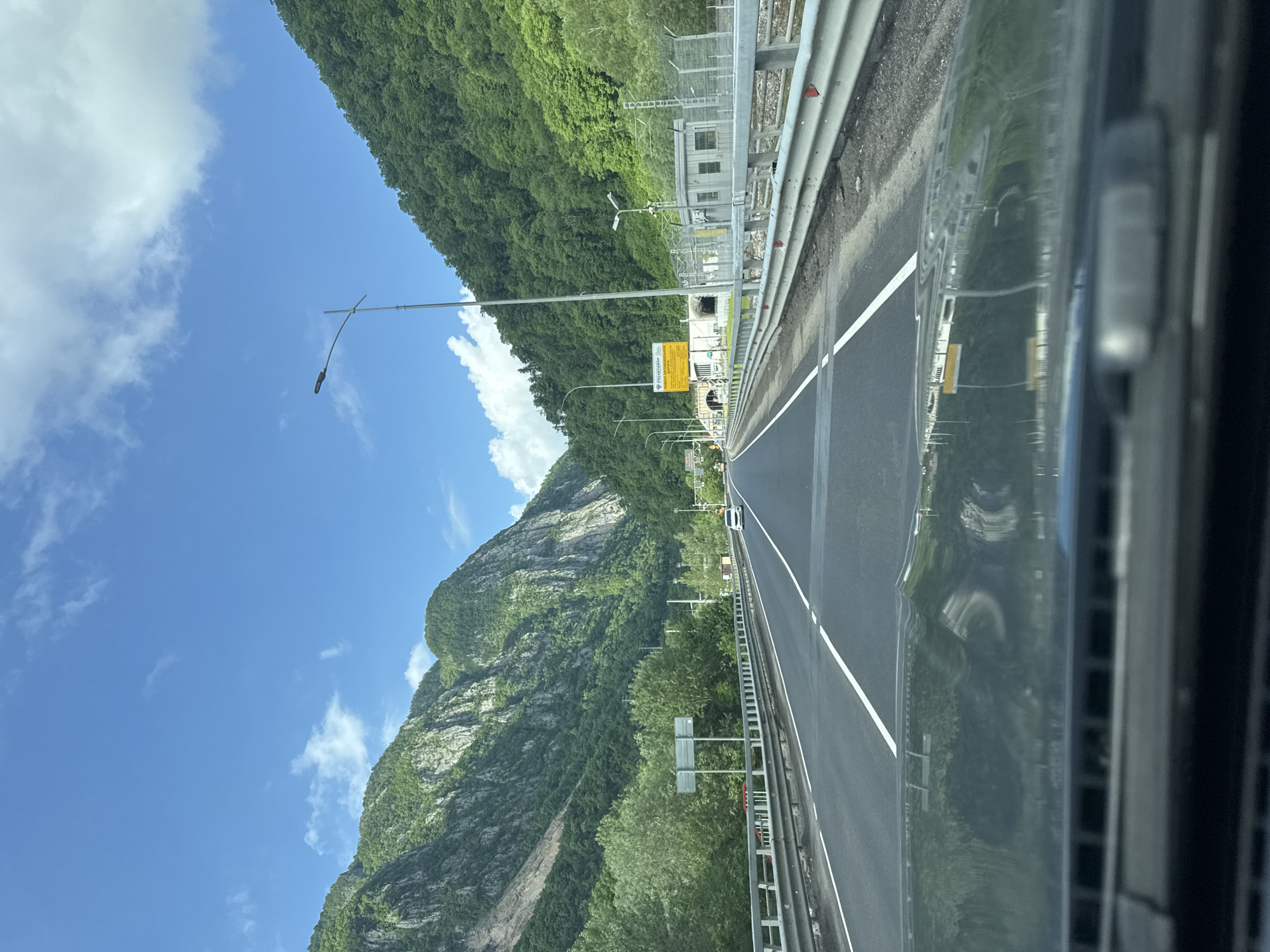 A highway traverses a mountainous landscape in Monastyr, Russia. The roadway, marked with white lines, is bordered by metal guardrails on both sides. A green, densely wooded mountain rises sharply on the left. The sky is blue with scattered white clouds. Streetlights are positioned along the road. In the distance, structures are visible adjacent to the road. The image appears to be taken from within a vehicle.