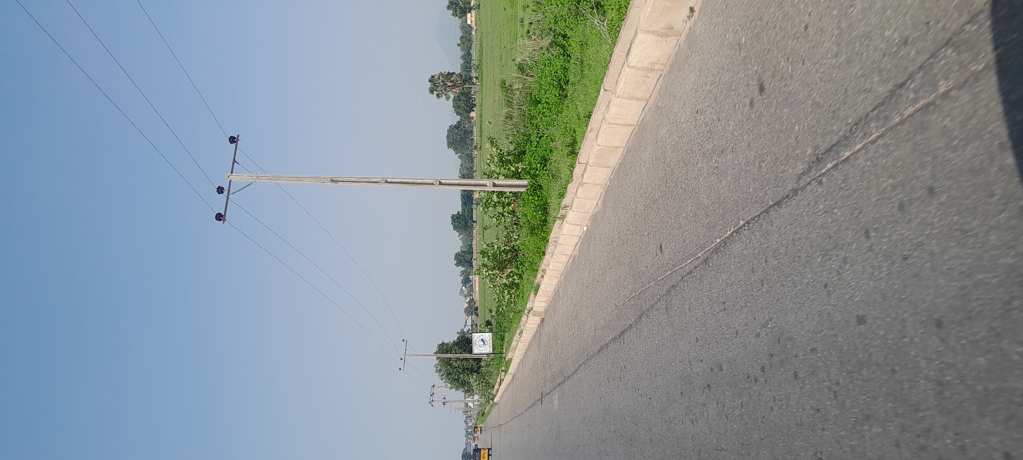 The image shows a roadside scene in Jalingo, Nigeria, under a clear blue sky. A paved road, with a visible lane marker, runs diagonally from the bottom left to the upper right. On the left side of the road, there are several yellow motorized rickshaws traveling in the same direction. Along the right side of the road, a concrete curb separates the pavement from a grassy area. Beyond the grass, a series of utility poles with power lines stretch into the distance. These poles are spaced along the side of the road, with one prominent pole in the foreground made of concrete. The background consists of a green field, dotted with trees, and some buildings in the far distance. A small white sign with a circular logo is visible on the left, near the curb. The overall impression is a rural or semi-rural highway setting.