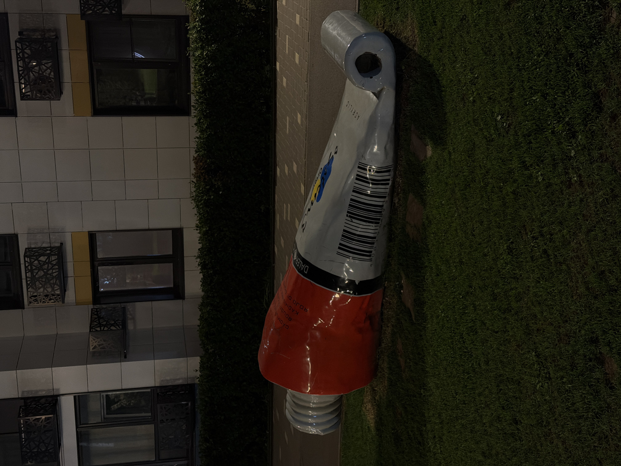 A nighttime outdoor shot captures a large, seemingly monumental sculpture of a squeezed paint tube, standing on a patch of grass near a building. The paint tube, with a predominantly red lower section, transitions to a grey upper section bearing what appears to be a barcode and partial text, including "ACRYLIC." The top of the tube is open, suggesting it's been used. The sculpture sits on green grass, beside a paved pathway. The building adjacent to the sculpture is modern, featuring dark windows and beige or grey exterior tiling. Dark greenery frames the scene on both sides, adding depth to the composition. The location appears to be Kazan, Russia, based on the markings on the paint tube sculpture.