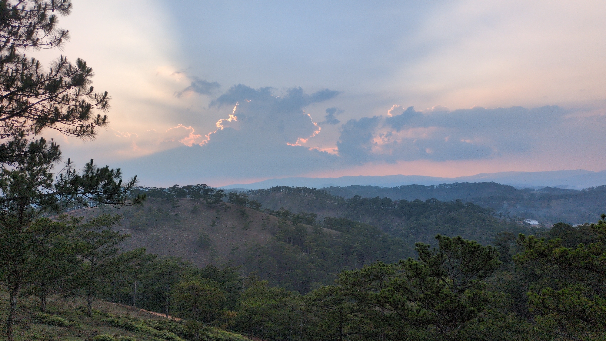 The image depicts a mountainous landscape in Lạc Dương, Vietnam, at twilight. The sky is a mix of blue, pink, and orange hues, with sunlight streaming through a layer of clouds. The foreground features pine trees, with a hillside covered in sparse vegetation. In the midground, a series of forested hills recede into the distance. A faint smoke plume can be seen rising from the landscape. Distant mountains appear as silhouettes against the sky.