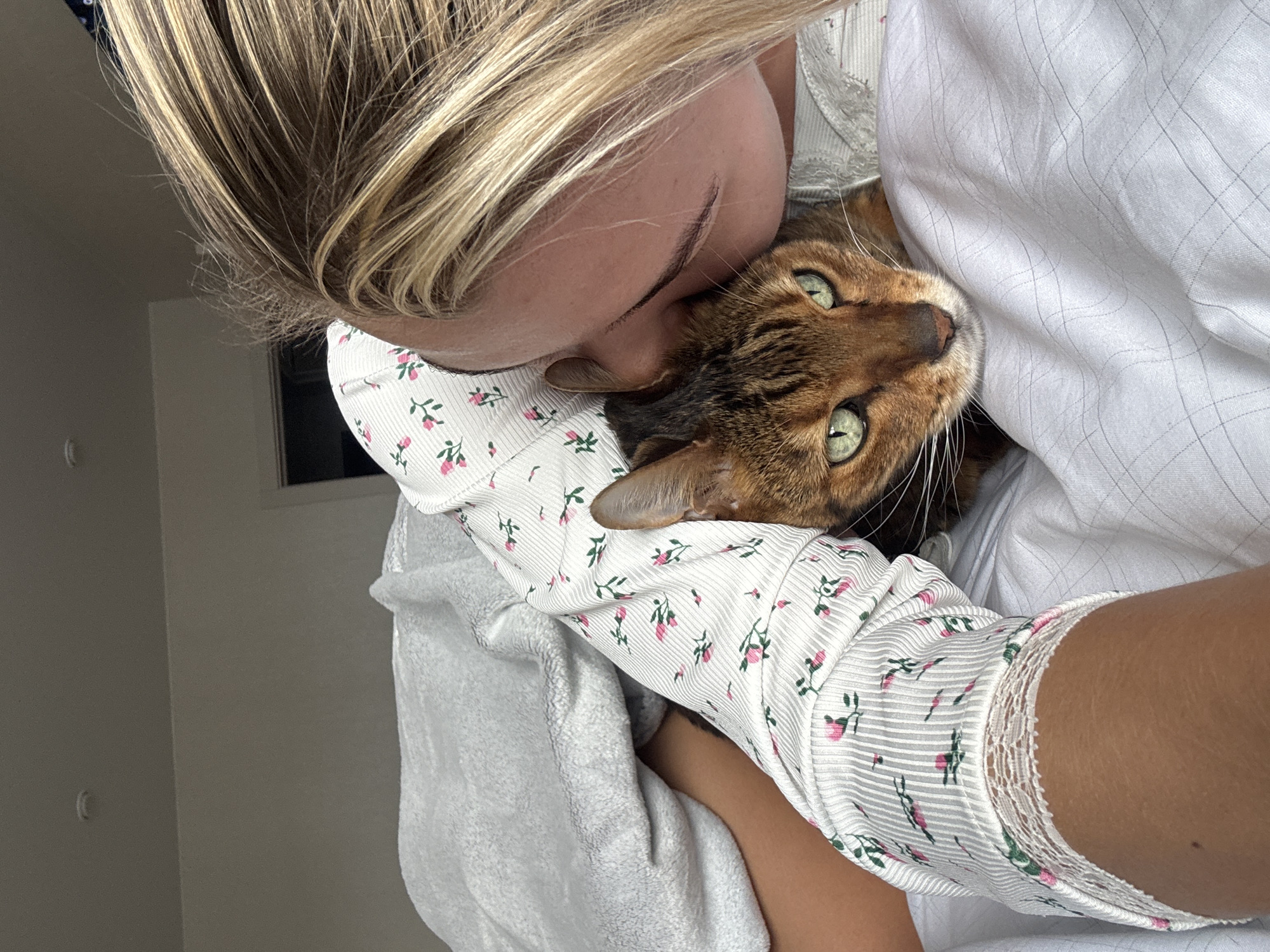 A woman with blonde hair is holding a Bengal cat. The woman is wearing a white, floral-patterned shirt with lace trim, and is embracing the cat closely, appearing to kiss its head. The cat is brown and black with green eyes. The background shows a white wall and a window, suggesting an indoor environment.