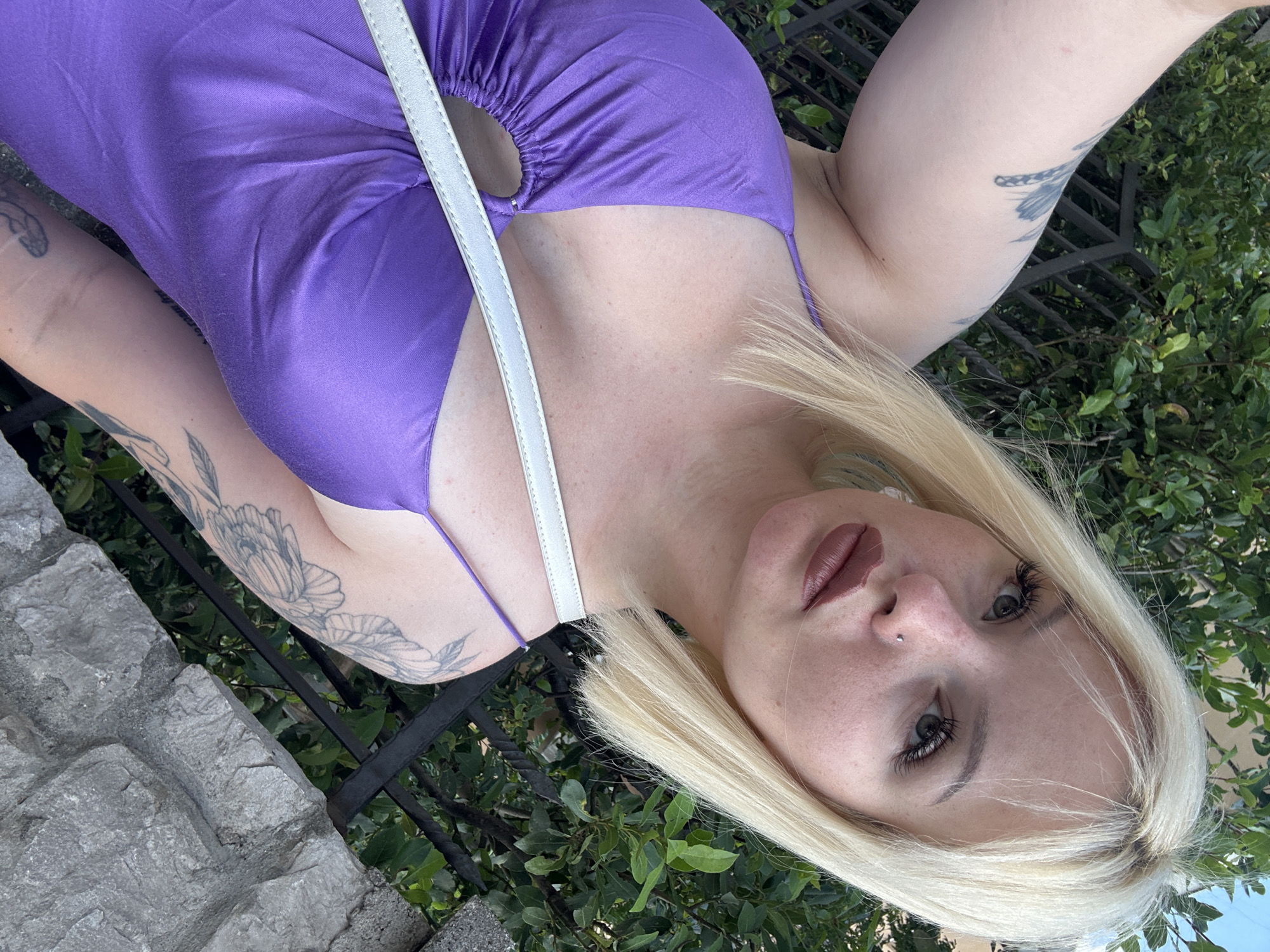 This is a portrait of a fair-skinned woman with blonde hair and dark grey eyes. She is wearing a vibrant purple satin dress with a halter neckline and a white strap across her chest. She has tattoos on her upper left arm and forearm. A small silver stud is visible on her nose. Her lips are painted with a matte, muted berry lipstick. The woman's head is tilted, and she is looking directly at the camera.
The background consists of lush green foliage and a dark metal fence. To the left of the frame, there is a textured stone surface, suggesting an outdoor setting. The lighting appears to be natural, implying daytime. The overall mood is casual and relaxed.
Based on the provided location context, this image was likely taken in Budva, Montenegro.