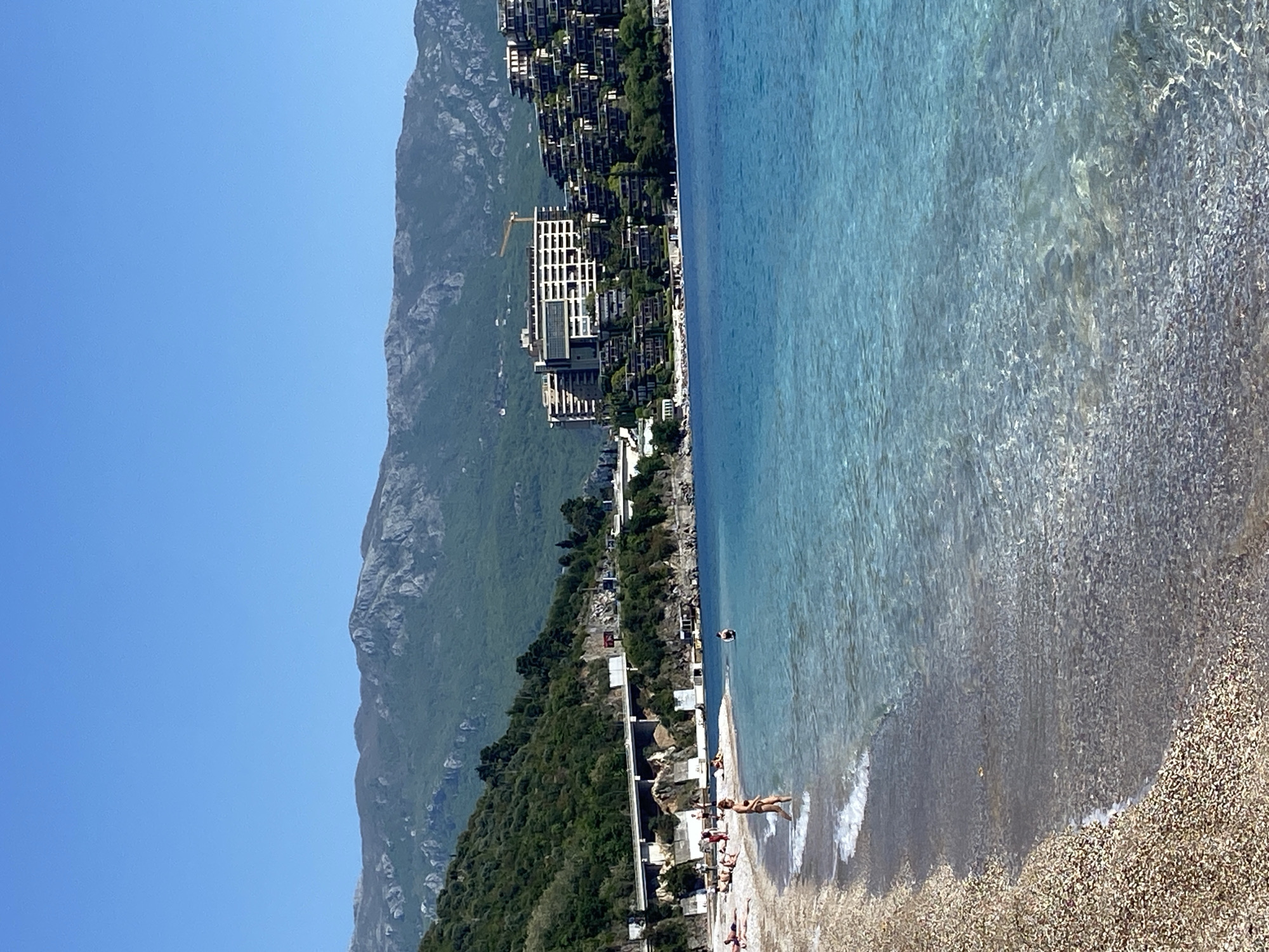 The image depicts a coastal scene in Budva, Montenegro, with a mountain range backdrop. A large building complex is visible along the coastline, with a construction crane present. The foreground features a beach with people sunbathing and standing near the water's edge. The water is clear and turquoise in color. The sky is clear and blue.