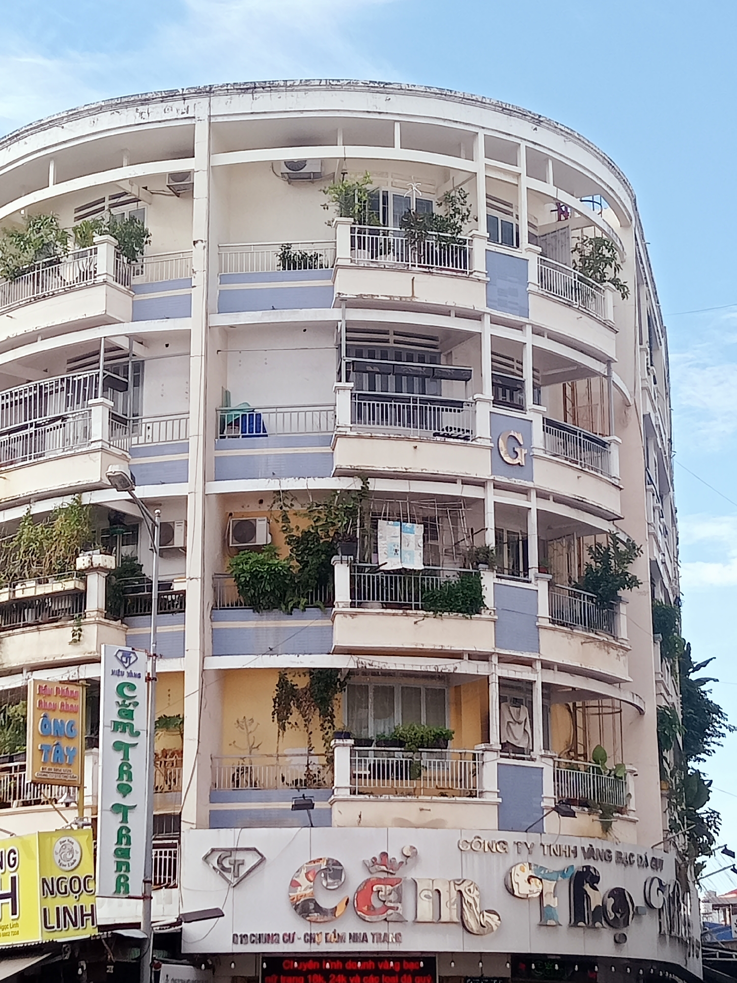 Here's a detailed description of the image:
The image shows a curved, multi-story building in Nha Trang Ward, Vietnam. The building appears to be a combination of residential and commercial spaces. The structure is mainly painted in white and blue with aging paint and signs of wear. The exterior of the building is adorned with multiple balconies with various railings and potted plants. Some of the balconies have clothes hanging out to dry. Air conditioning units are visible on some of the walls.
The ground floor of the building features commercial signage. Visible signage includes the text "Công Ty Tnhh Vàng Bạc Đá Qui," and "CentToy," presumably indicating a jewelry or gem-related business. Other nearby business signs are partially visible on the left side of the building.
The overall impression is that the building is in an urban setting, likely in a busy commercial district within Nha Trang Ward, Vietnam. The architectural style is somewhat dated and shows the effects of time and environmental exposure.