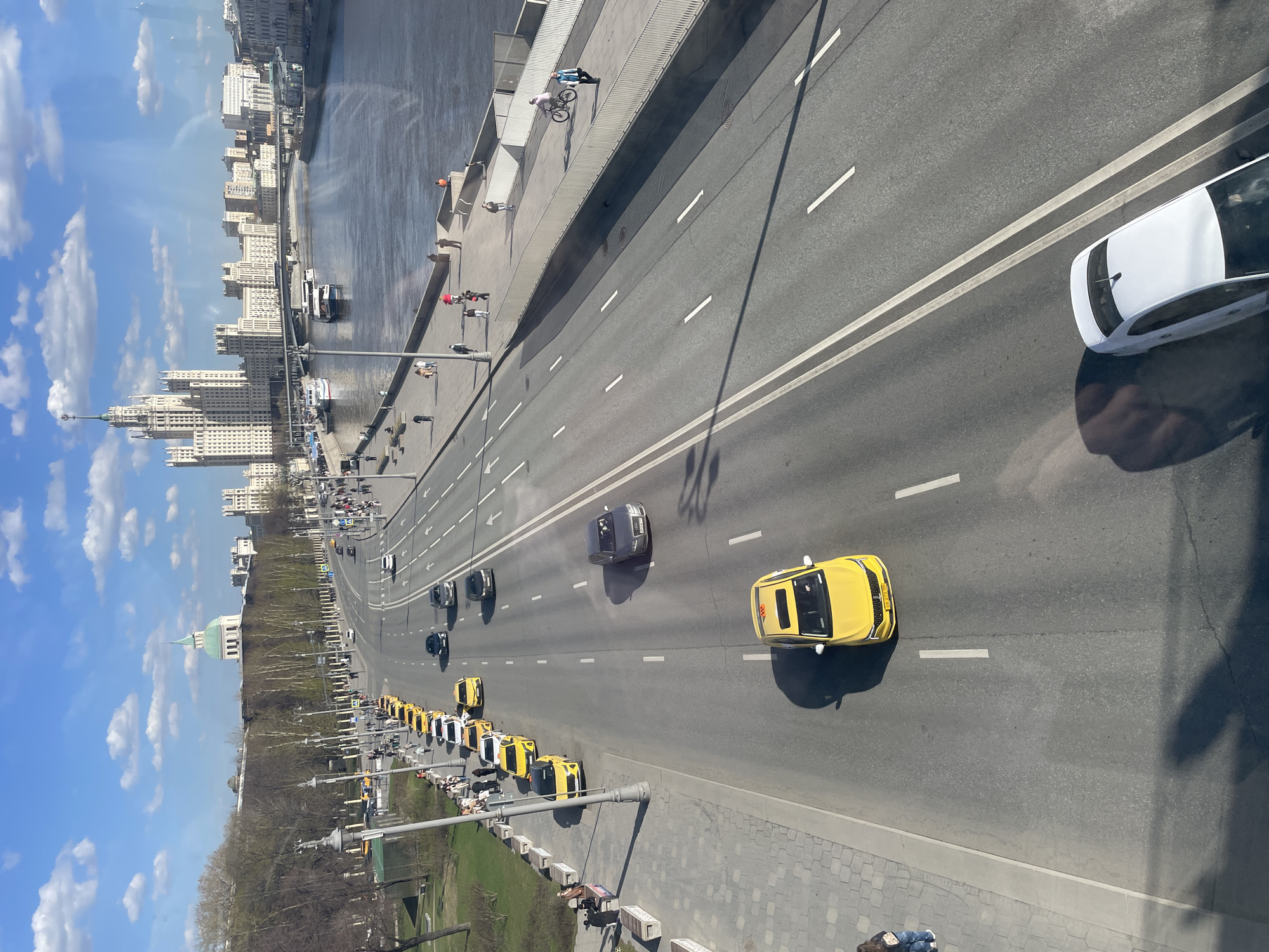 The image captures an elevated view of a Moscow streetscape. A multi-lane asphalt roadway dominates the central part of the frame, populated with vehicular traffic including numerous yellow taxis. A white car is visible at the top right. The image includes pedestrian areas lined with benches and trees along the sides of the road. A wide river runs parallel to the road, with various buildings and cityscape elements visible in the distance on the opposite bank. The sky is partly cloudy. A cyclist is visible on the street. The composition suggests the image was captured from a high vantage point, such as an upper floor window, with the reflection of the photographer's hand visible on the window.