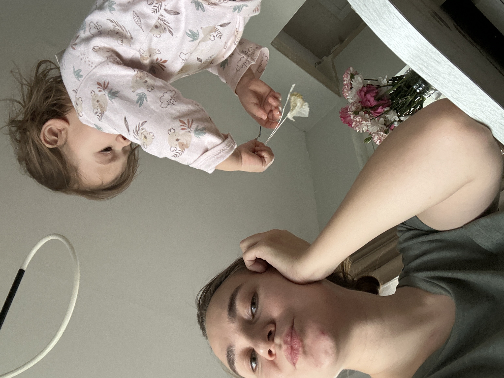 A woman is pictured, looking downwards at the camera. She is wearing a grey t-shirt. A child, possibly a toddler, wearing pink pajamas with a koala bear pattern, is above her, reaching for a bouquet of pink and white flowers that are on a white, possibly reflective, surface. A white, curved object is visible in the lower left corner. The location is not discernable from the image, so the information about Kharnavy, Russia, cannot be verified.