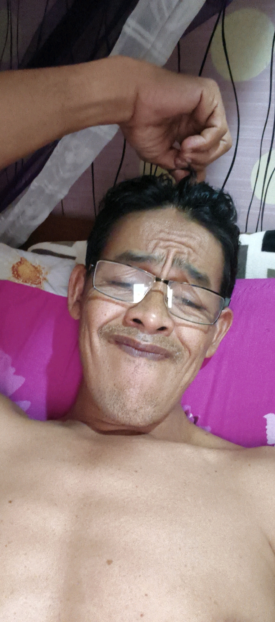 An adult male is lying down and looking at the camera, with his bare chest and shoulders visible. He is wearing glasses and has a mustache. His left arm is raised and bent, with his hand near his head. The background includes a pink surface, possibly a pillow or blanket, and a patterned fabric. The image appears to be a selfie taken indoors. The location of Muaro Jambi, Indonesia is not depicted or inferable from the image.